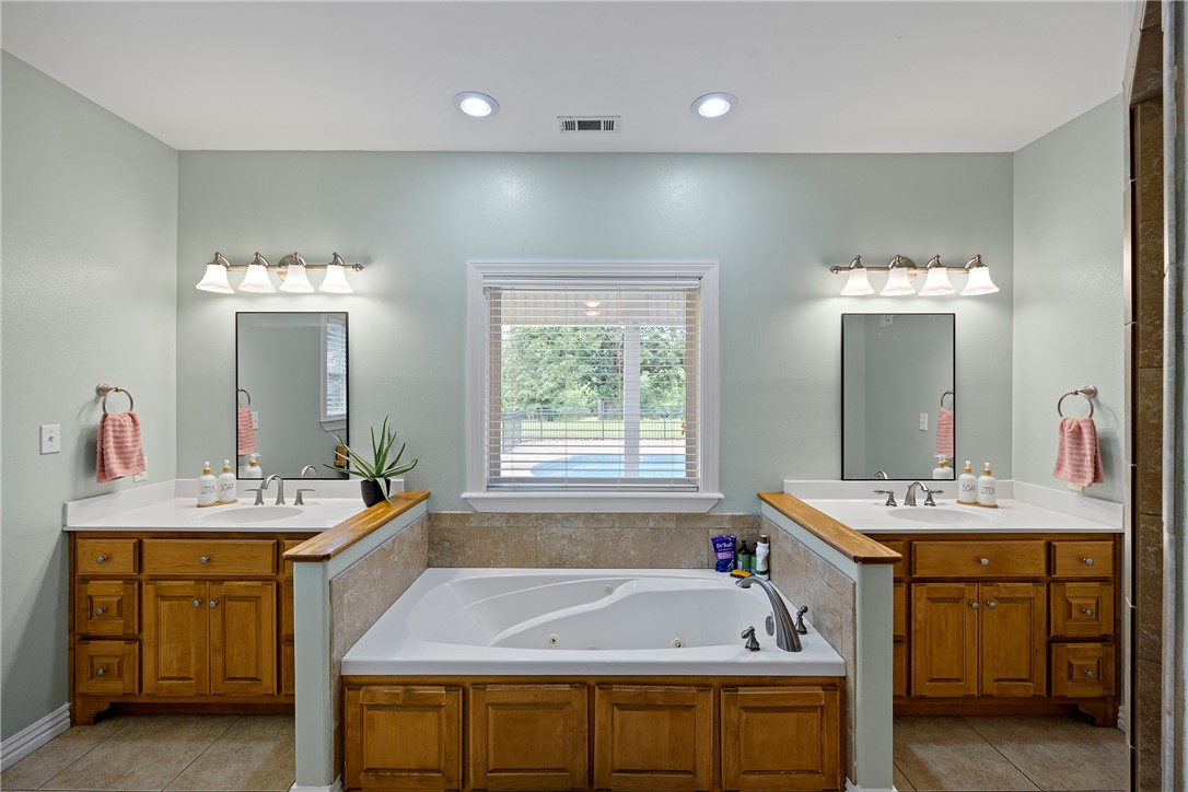 751 Walthall Road Russellville, AR 72802 - Photo 18 of 60 Soaking tub with tile surround, flanked by two wood-finish vanities featuring white countertops, undermount sinks, and wall-mounted light fixtures