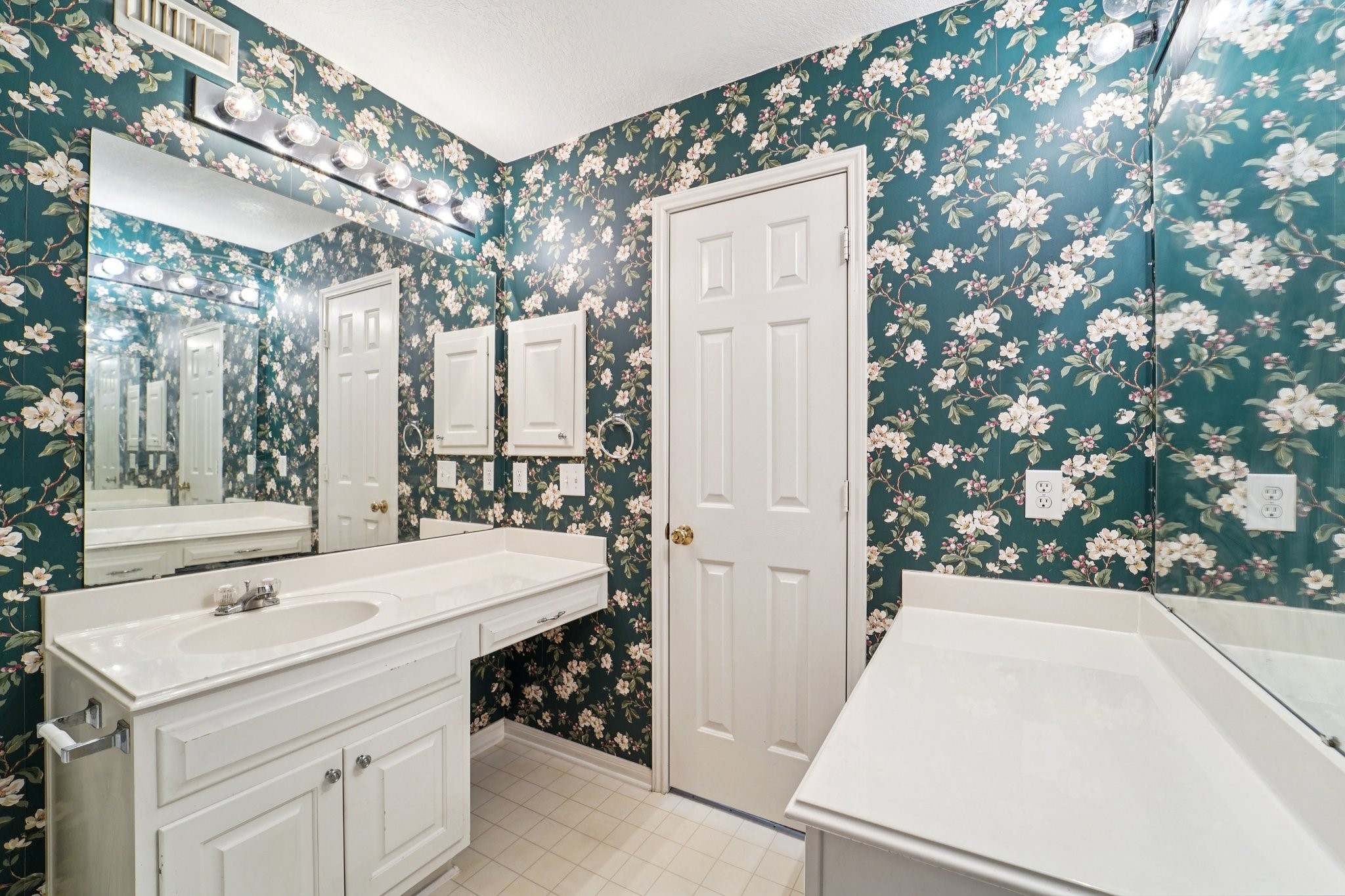 14420 Walters Road, Unit 55 Houston, TX 77014 - Photo 30 of 35 This bathroom features floral wallpaper, a large mirror with vanity lighting, a white countertop with ample storage, and a white door.