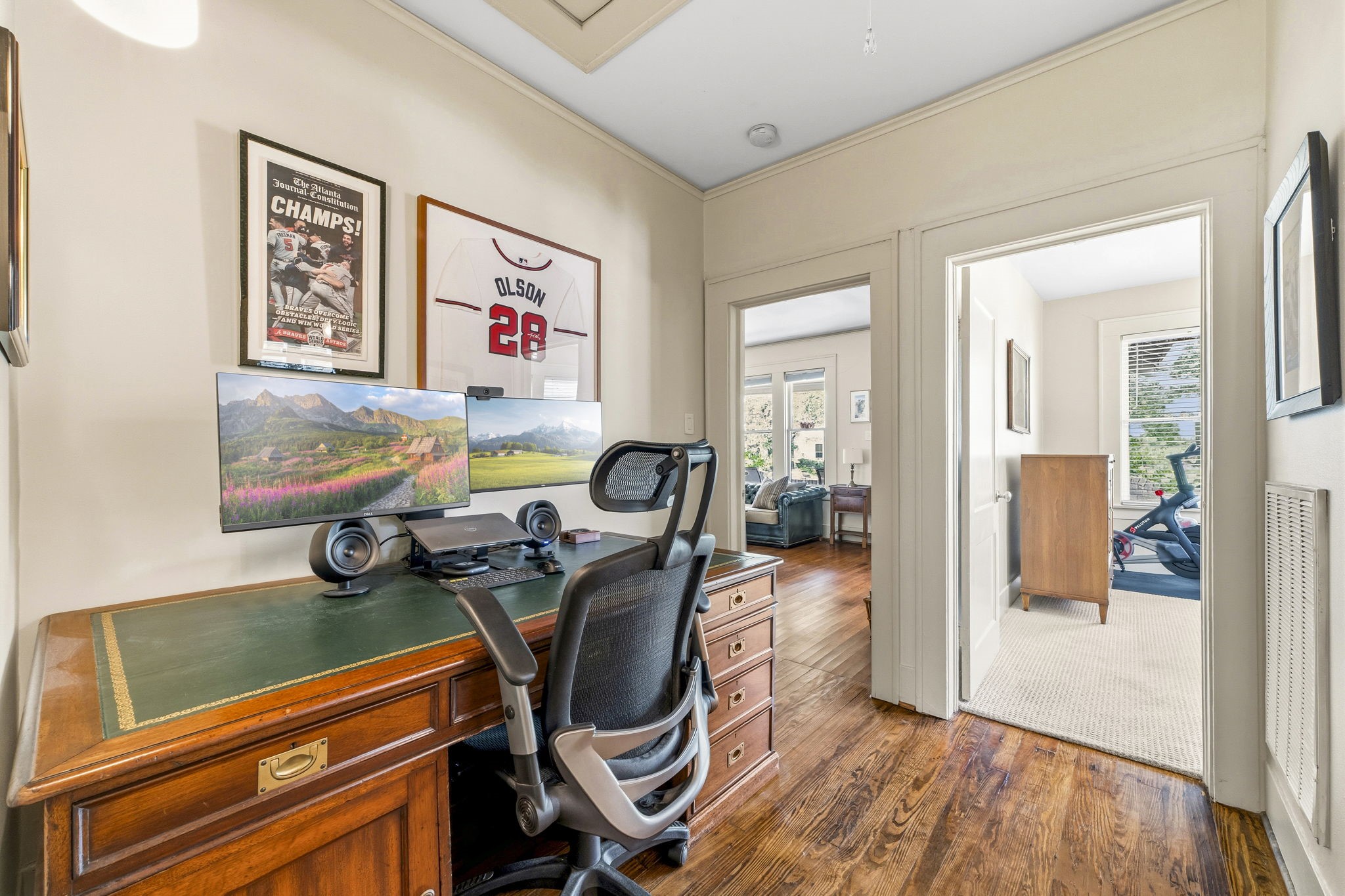 1521 Kipling Street Houston, TX 77006 - Photo 28 of 35 This photo showcases a cozy home office with hardwood floors and natural light. It features a classic wooden desk, dual monitors, and sports-themed wall decor. Adjacent rooms include a living area and an exercise space with a stationary bike.