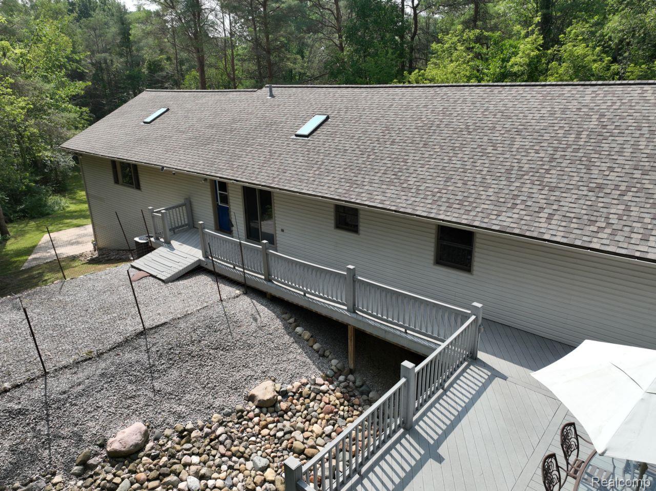 10201 Washburn Road Goodrich, MI 48438 - Photo 72 of 85 Light-toned siding exterior featuring a composite shingle roof with skylights, a multi-level deck with gray railings, and a gravel hardscaped area with river rock accents