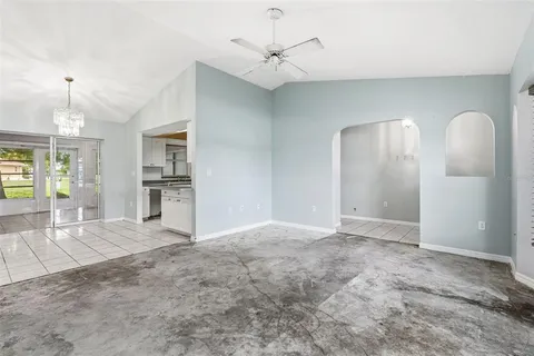 an empty room with chandelier fan and kitchen view