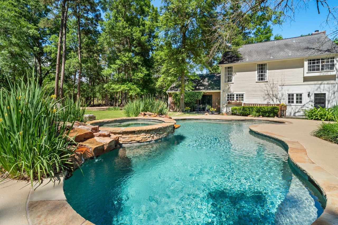 36 Wind River Drive Conroe, TX 77384 - Photo 13 of 45 This photo features a charming two-story home with a spacious backyard, highlighted by a beautiful swimming pool and spa surrounded by lush greenery. The setting is tranquil, with mature trees providing privacy and a serene atmosphere. Perfect for relaxation and outdoor entertaining.
