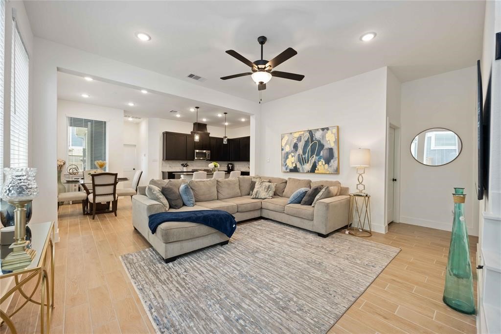 10014 Nectar Path Iowa Colony, TX 77583 - Photo 5 of 26 This inviting open-concept living area features a spacious sectional sofa, modern decor, and a ceiling fan. The room seamlessly connects to a dining area and a sleek kitchen with dark cabinetry and stainless steel appliances, all under warm recessed lighting.