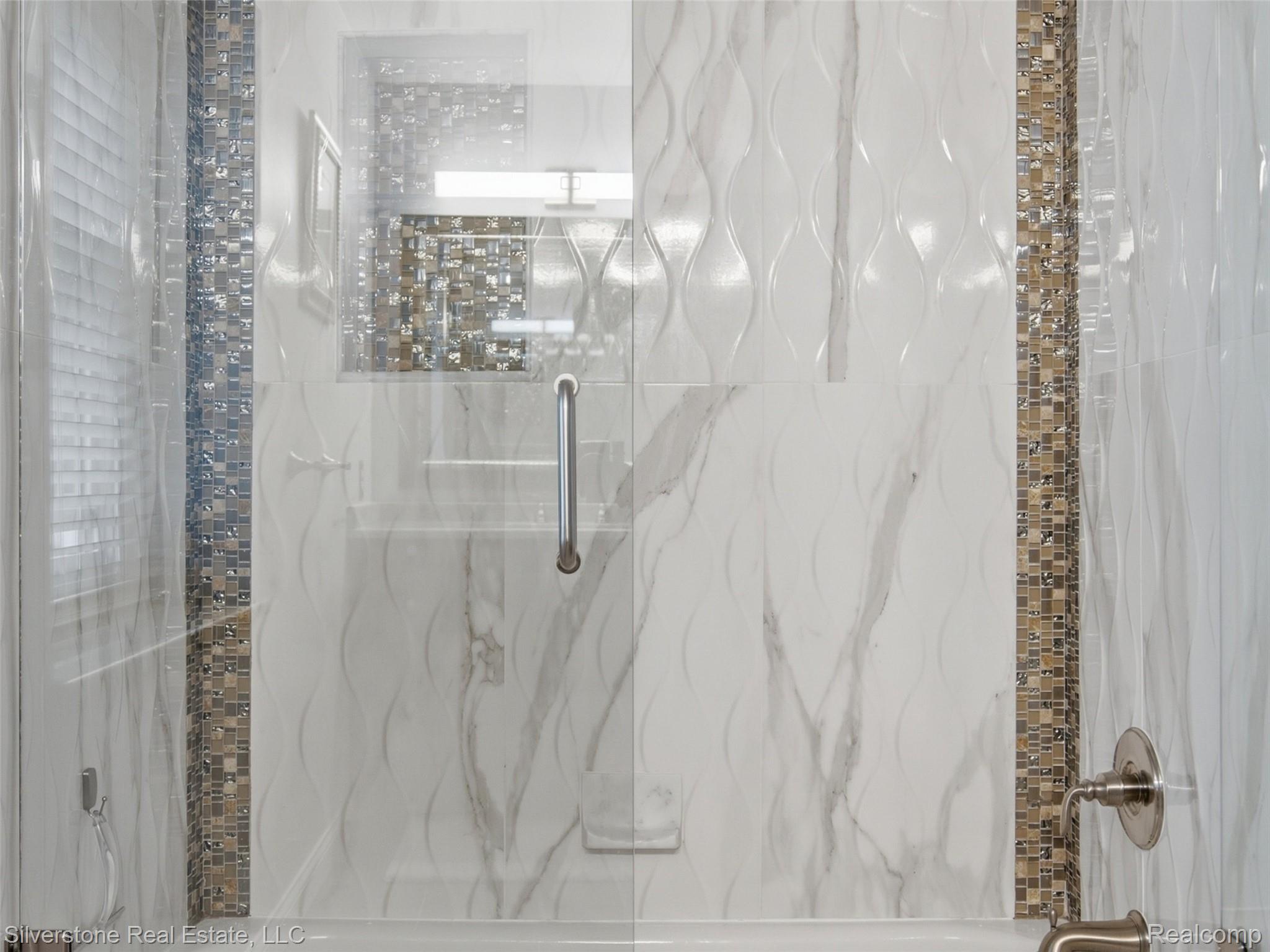 6577 Edith Court Troy, MI 48085 - Photo 43 of 61 Shower enclosure featuring a clear glass door with a brushed nickel handle, surrounded by white marble-look tile with wave-patterned accent tiles, and a mosaic tile vertical accent strip