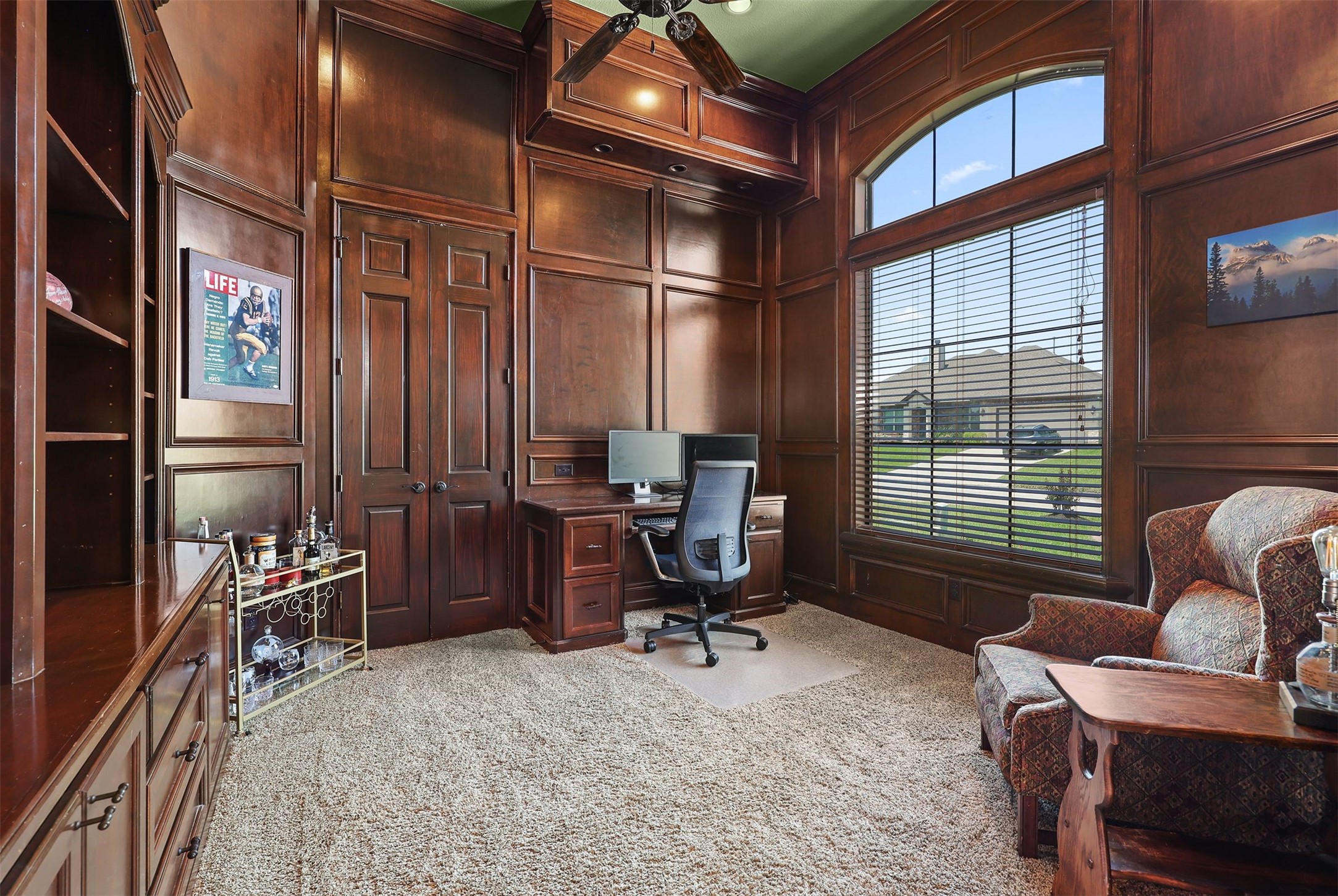 10896 Decatur Street Willis, TX 77318 - Photo 13 of 50 This elegant home office, adorned with rich wood paneling and built-in shelving, boasts a large window with blinds for ample natural light, complemented by a comfortable chair, computer desk, and cozy seating area, creating an inviting and productive workspace.