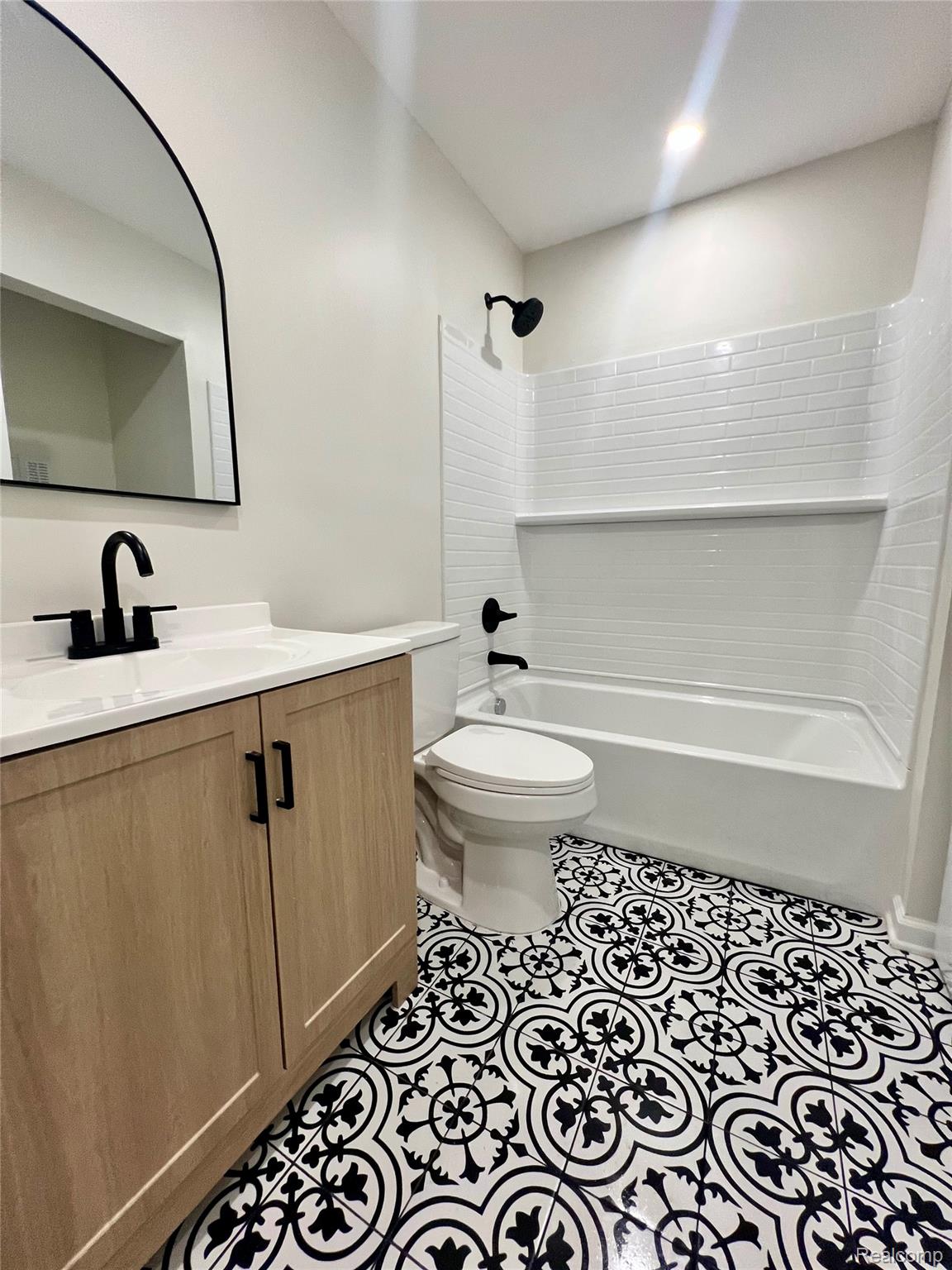 219 O'Doherty Road Brighton, MI 48116 - Photo 15 of 18 Bathroom featuring patterned floor tiles, light wood-finish vanity with black hardware, white countertop, black faucet, and a black-framed arched mirror