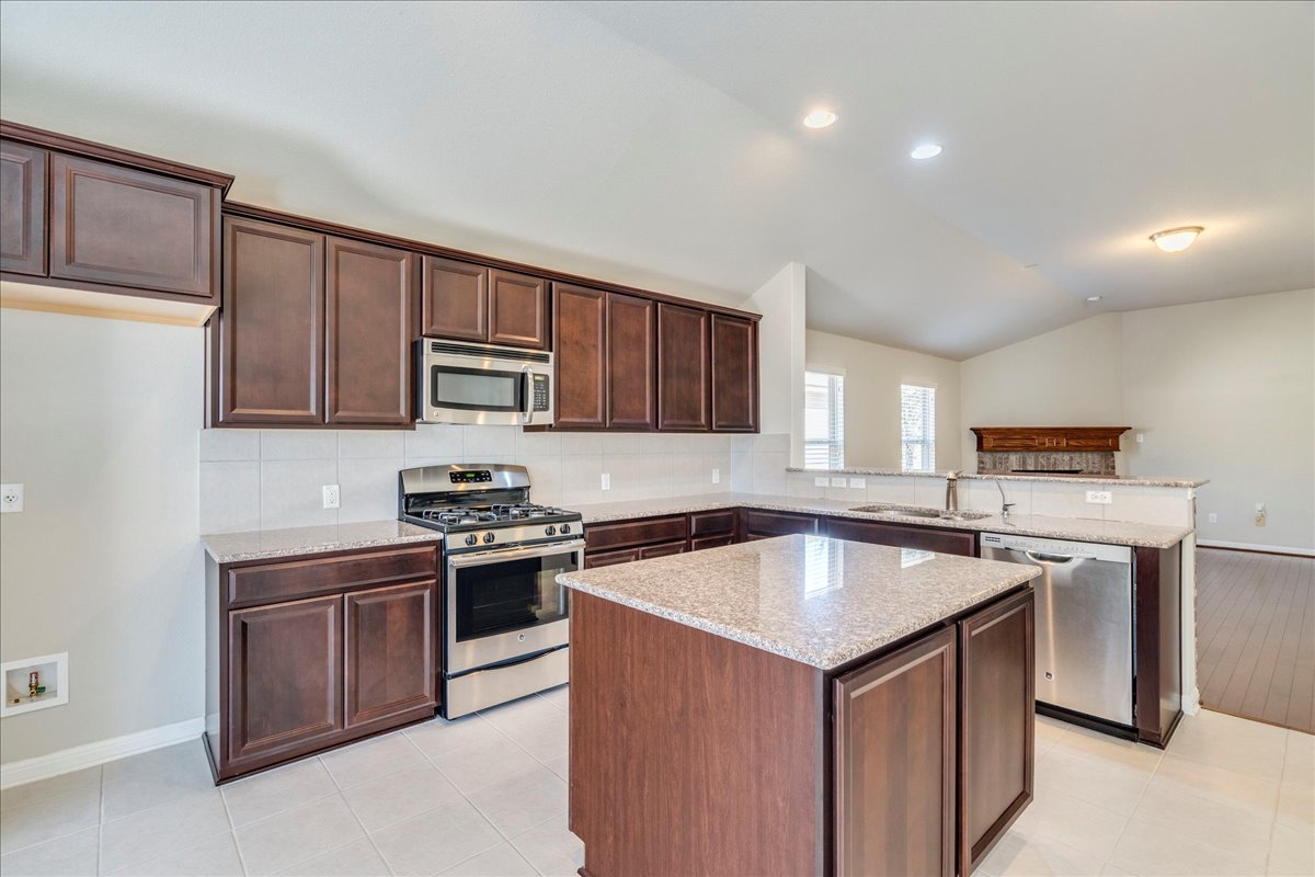 161 Mistflower Lane Kyle, TX 78640 - Photo 11 of 37 Kitchen featuring stainless steel appliances, dark wood finish cabinetry, a kitchen island, light stone countertops, and a peninsula