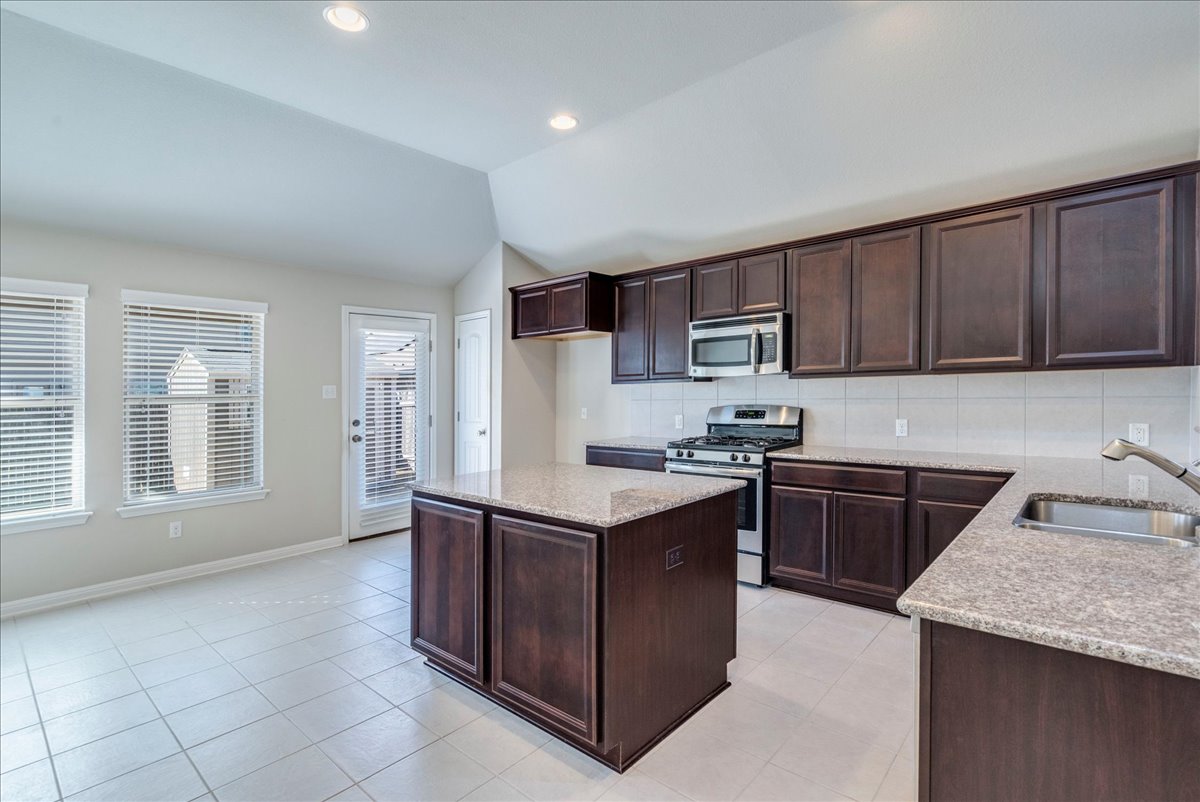 161 Mistflower Lane Kyle, TX 78640 - Photo 9 of 37 Kitchen with dark wood finish cabinets, stainless steel appliances, a kitchen island, vaulted ceiling, and light stone counters