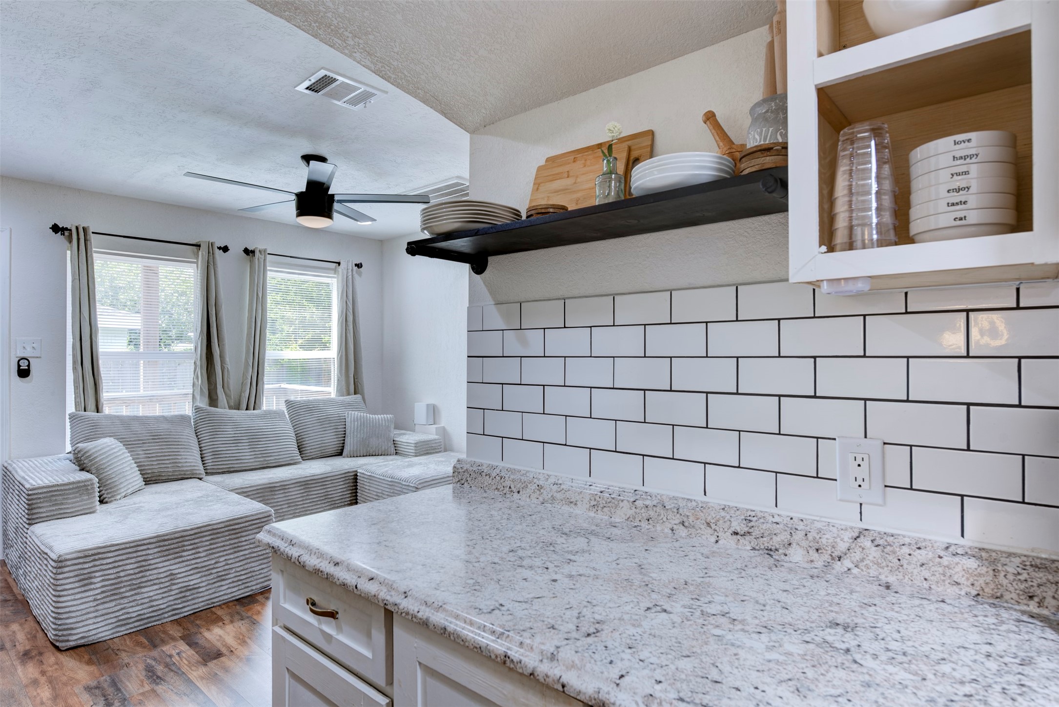 1121 Panama Street Houston, TX 77009 - Photo 9 of 35 Kitchen pass-through view showing granite countertops with white and gray veining, white subway tile backsplash with dark grout, black steel open shelving with pipe brackets, white shaker cabinetry with brushed gold hardware, ceiling fan with light kit, and oversized window with grommet curtain panels.