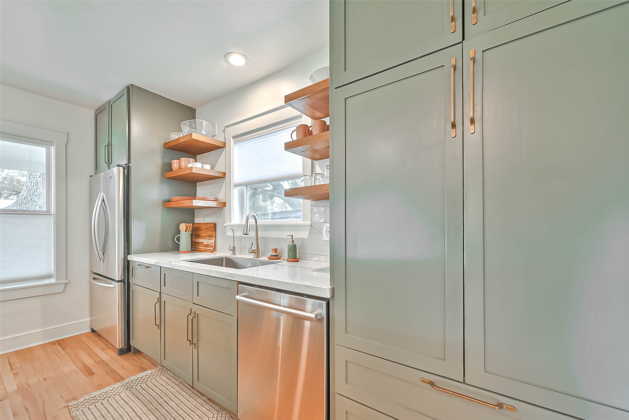 412 Sue Street Houston, TX 77009 - Photo 3 of 19 This kitchen features sleek light green cabinetry with gold hardware, a stainless steel fridge and dishwasher, and open wooden shelves for added warmth. There's ample natural light from the window above the sink, and the room has a cozy, modern feel with light wood flooring. Open to the living/dining area for entertaining.