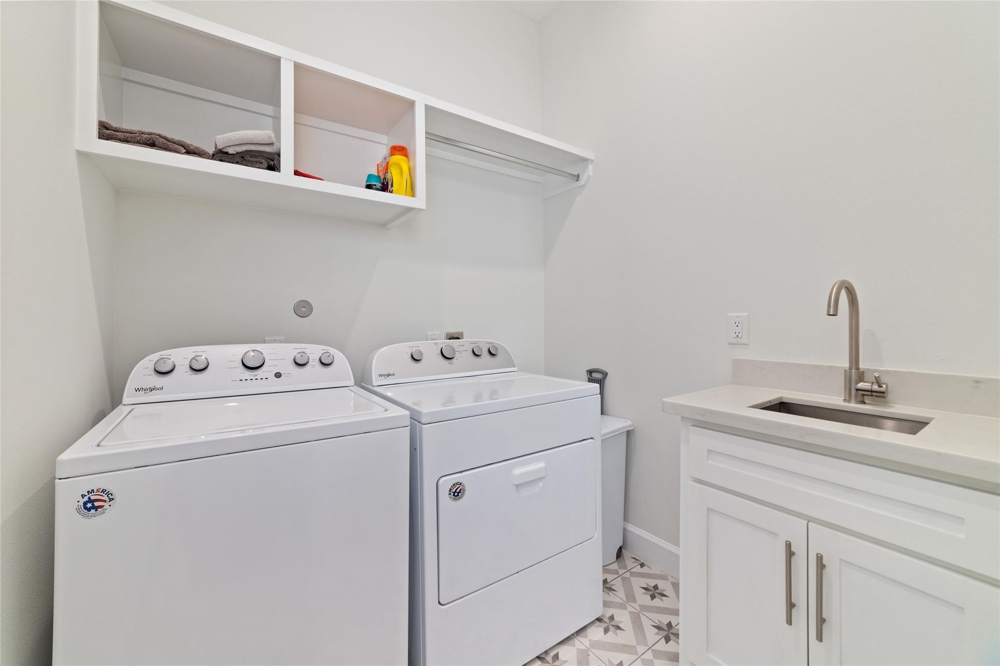 710 Delano Street, Unit A Houston, TX 77003 - Photo 24 of 32 This is a bright, modern laundry room featuring a washer and dryer set, a sink with a countertop, and open shelving for storage. The patterned tile floor adds a stylish touch.