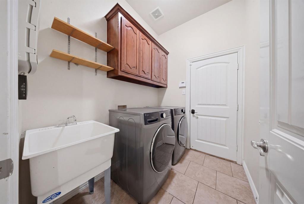 603 Cedar Drive Pilot Point, TX 76258 - Photo 29 of 39 Utility room featuring a built-in utility sink, overhead wooden cabinetry, open shelving, and tiled flooring