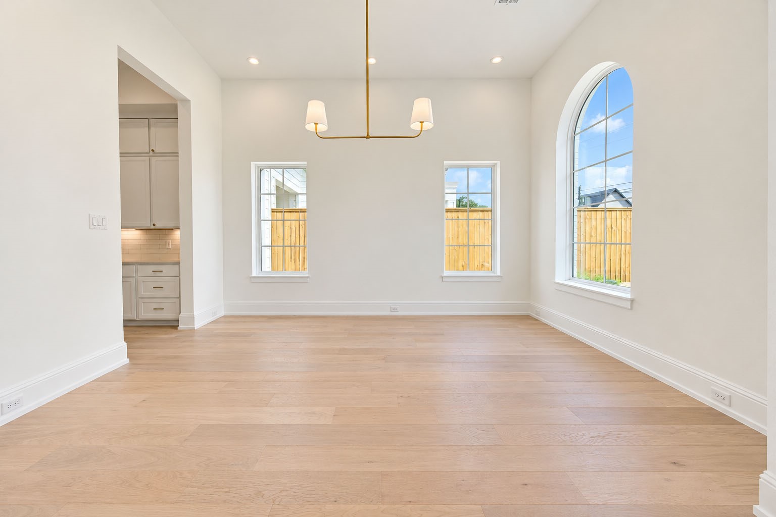 2309 Hollister Road Houston, TX 77080 - Photo 7 of 47 Another view of the elegant dining room highlights its generous proportions, soaring ceiling, warm wide-plank flooring, and refined statement lighting. A dramatic arched window paired with additional windows bathes the room in natural light, while the open connection to the kitchen creates an effortless setting for sophisticated dinner parties, holiday celebrations, and everyday entertaining.