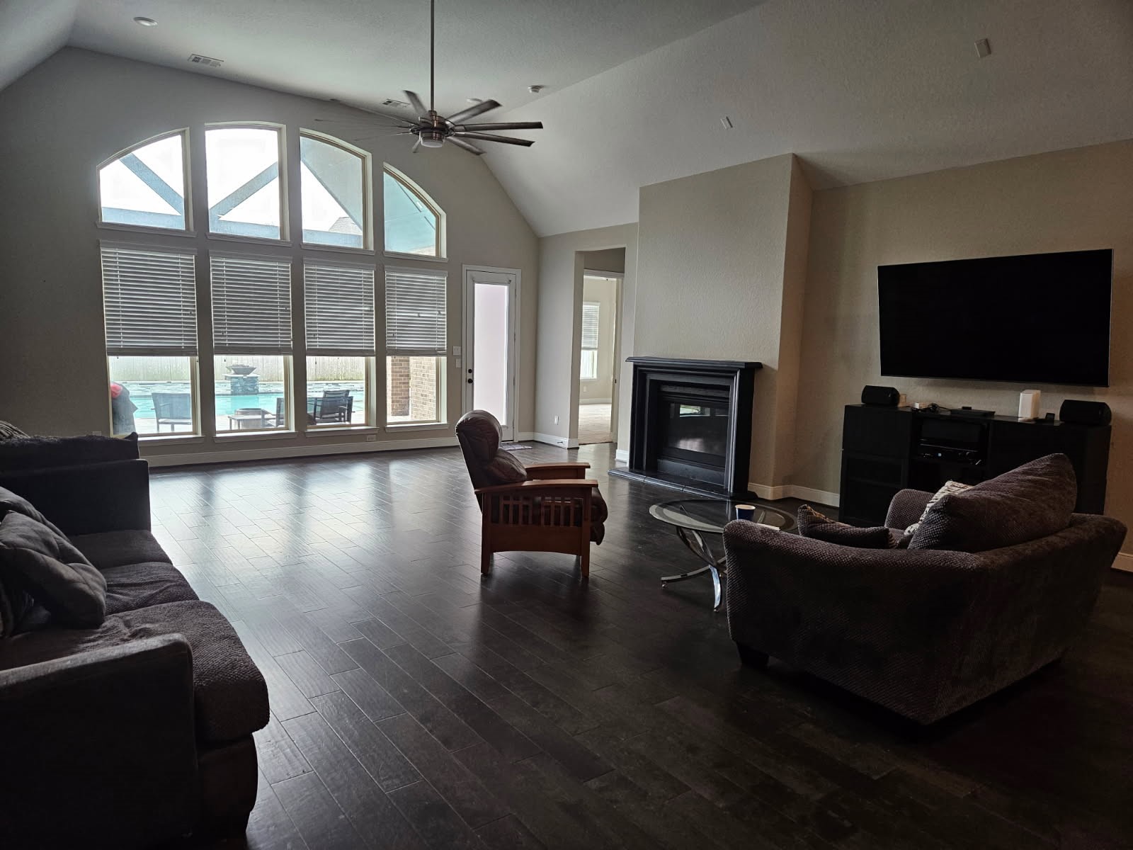 18919 Steep Step Trail Cypress, TX 77433 - Photo 2 of 12 Spacious living room with high ceilings, dark wood flooring, and large arched windows offering a view of the pool. Features a fireplace, ceiling fan, and wall-mounted TV, creating a cozy yet modern atmosphere.