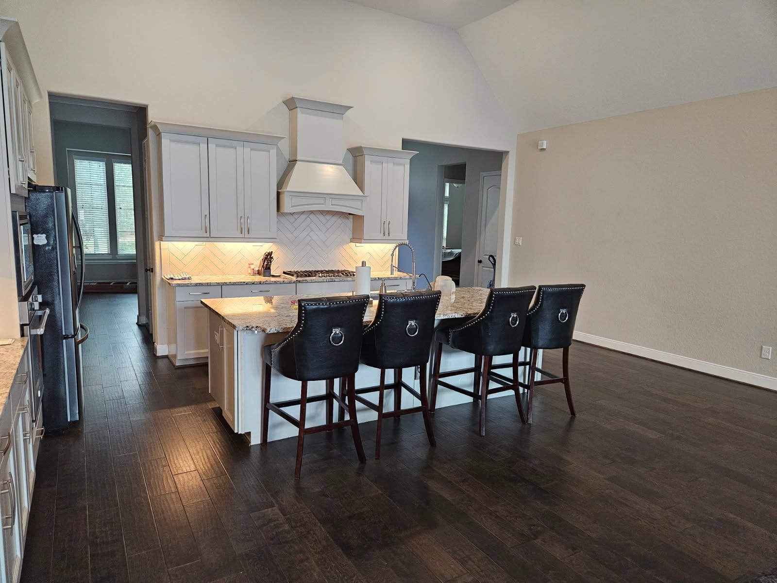 18919 Steep Step Trail Cypress, TX 77433 - Photo 6 of 12 This photo shows a spacious kitchen with dark hardwood floors, an island with barstools, and white cabinetry. The kitchen features a modern range hood, granite countertops, and a herringbone backsplash, providing a stylish and functional cooking space.