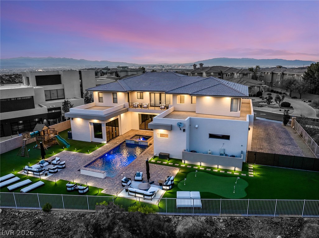 5 Crystal Tree Pass Henderson, NV 89052 - Photo 62 of 67 An opulent, modern home showcases an illuminated pool, putting green, and playset beneath a vibrant sunset.