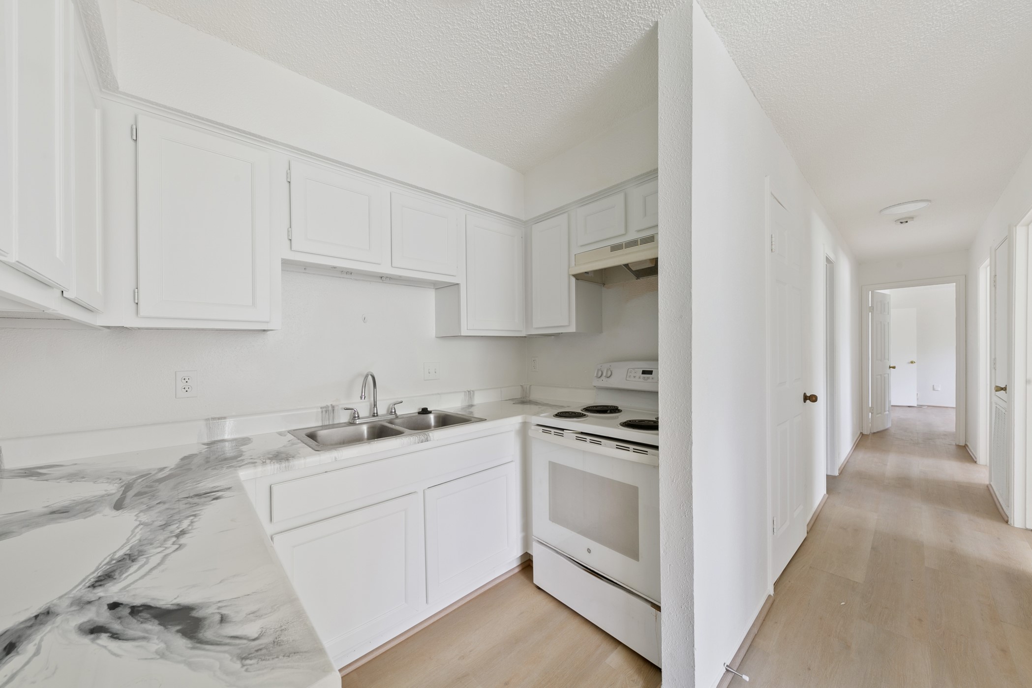 700 Lyle W Avenue, Unit D Cleveland, TX 77327 - Photo 5 of 16 This photo features a bright, compact kitchen with white cabinetry and appliances, and a marbled countertop. It opens to a hallway with light wood flooring leading to other rooms, creating a clean and airy feel.
