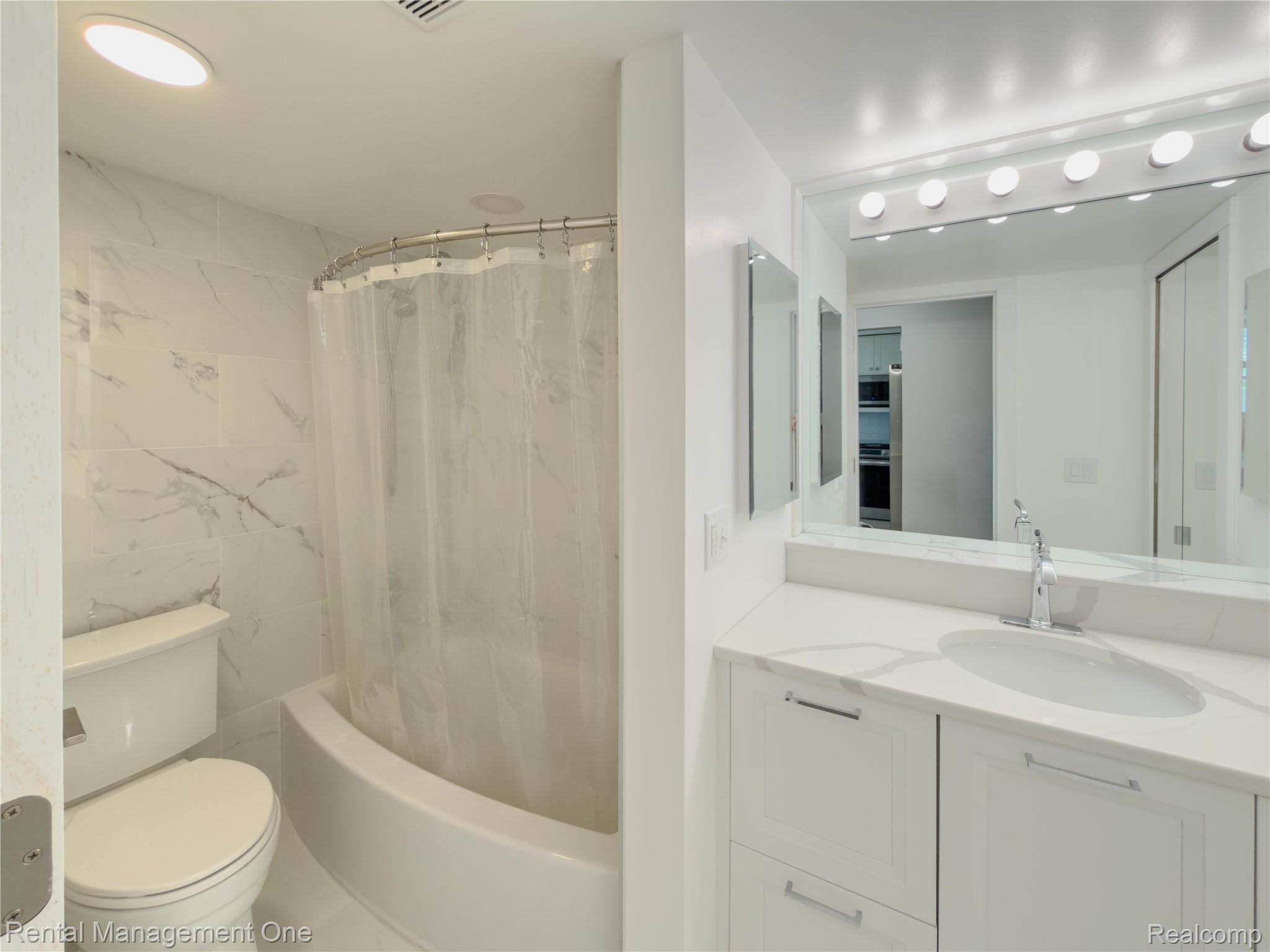 1603 Cass Lake Road, Unit D Keego Harbor, MI 48320 - Photo 9 of 20 Modern bathroom featuring a white vanity with an integrated sink, a large mirror with Hollywood-style lighting, and a bathtub with a curved shower rod