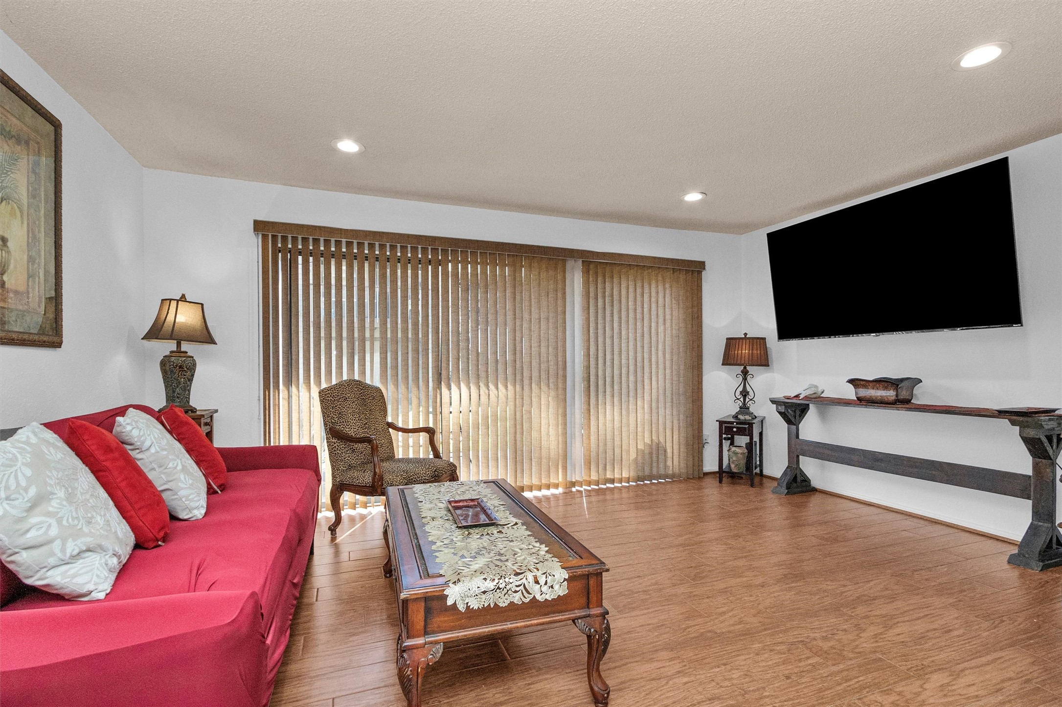 12900 Walden Road, Unit D 411 Montgomery, TX 77356 - Photo 6 of 18 Step into a welcoming living room featuring warm wood floors and abundant natural light through large windows with vertical blinds, complemented by recessed lighting. Relax on the cozy red sofa with accent pillows, or the classic armchair, while enjoying a movie on the wall-mounted flat-screen TV. The space is elegantly finished with a wooden coffee table adorned with decorative lace and stylish side tables with lamps.