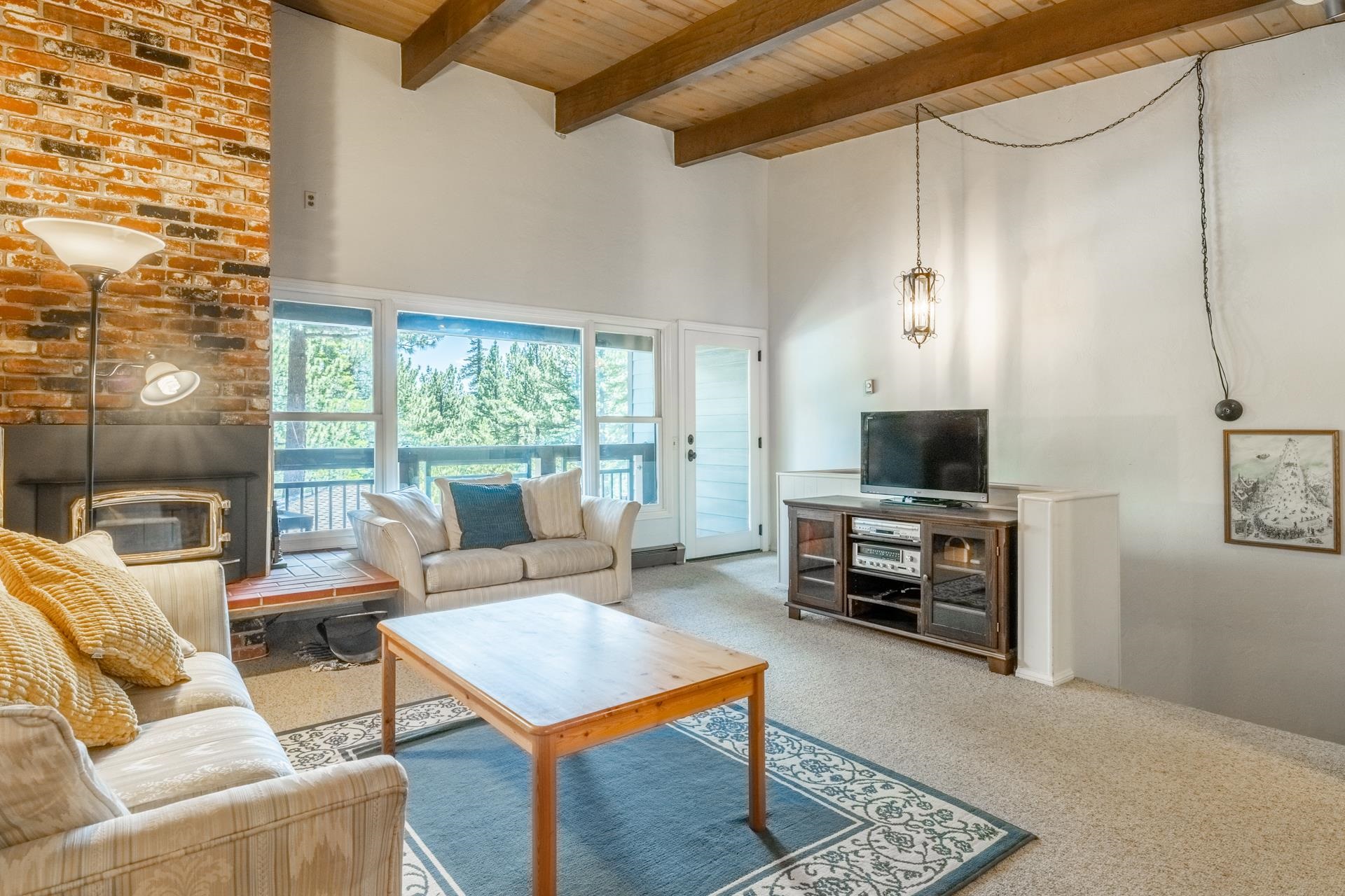 50 Hidden Valley Road, Unit 17 Mammoth Lakes, CA 93546 - Photo 8 of 40 Living space featuring a natural brick fireplace, a wood-burning stove, exposed wood beam ceilings, and large windows overlooking exterior greenery