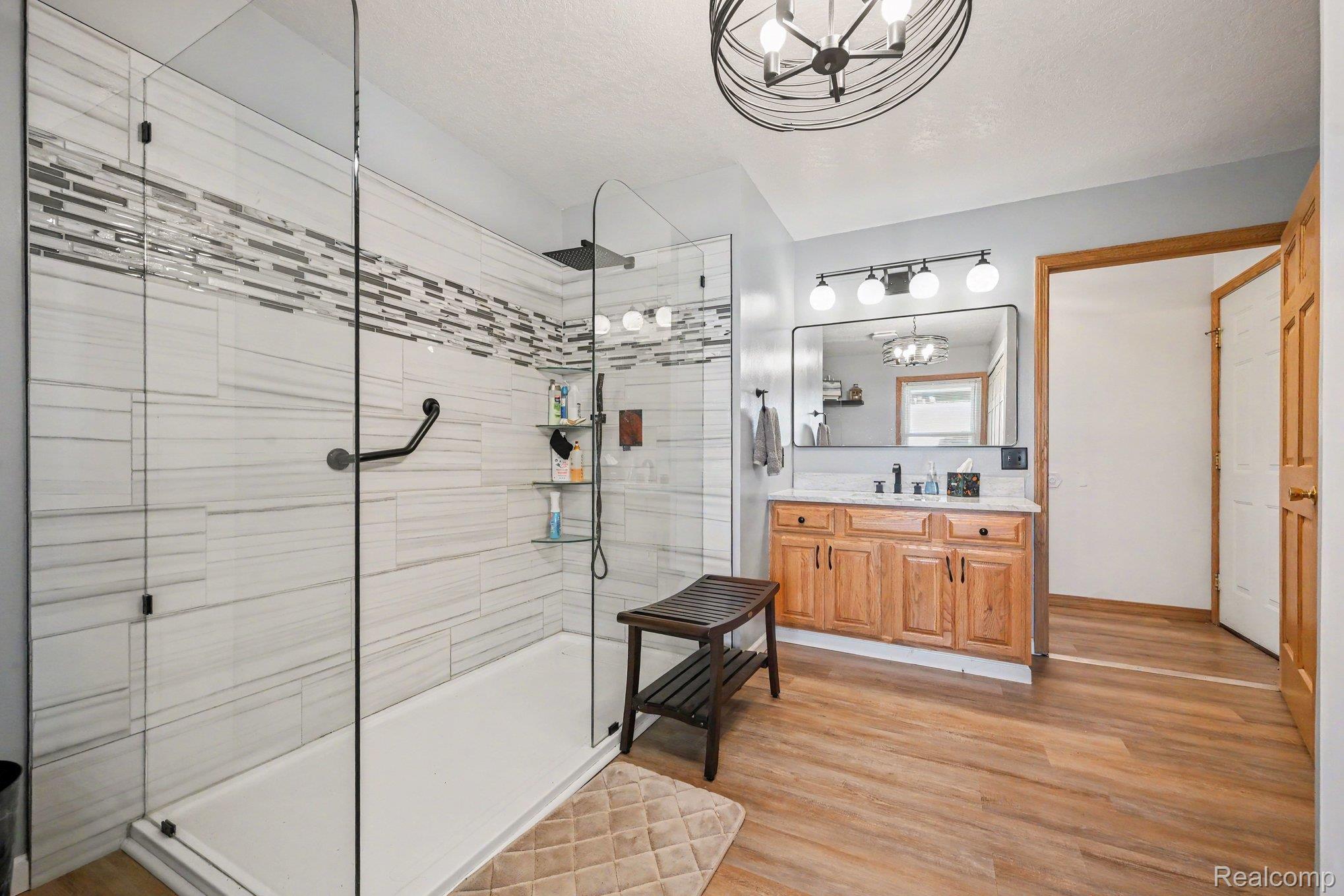 996 Arbogast Road Union City, MI 49094 - Photo 14 of 34 Spacious bathroom featuring a glass-enclosed shower with white and grey horizontal tile, a black ceiling light fixture, a wood-finish vanity with a white countertop, and wood-finish flooring