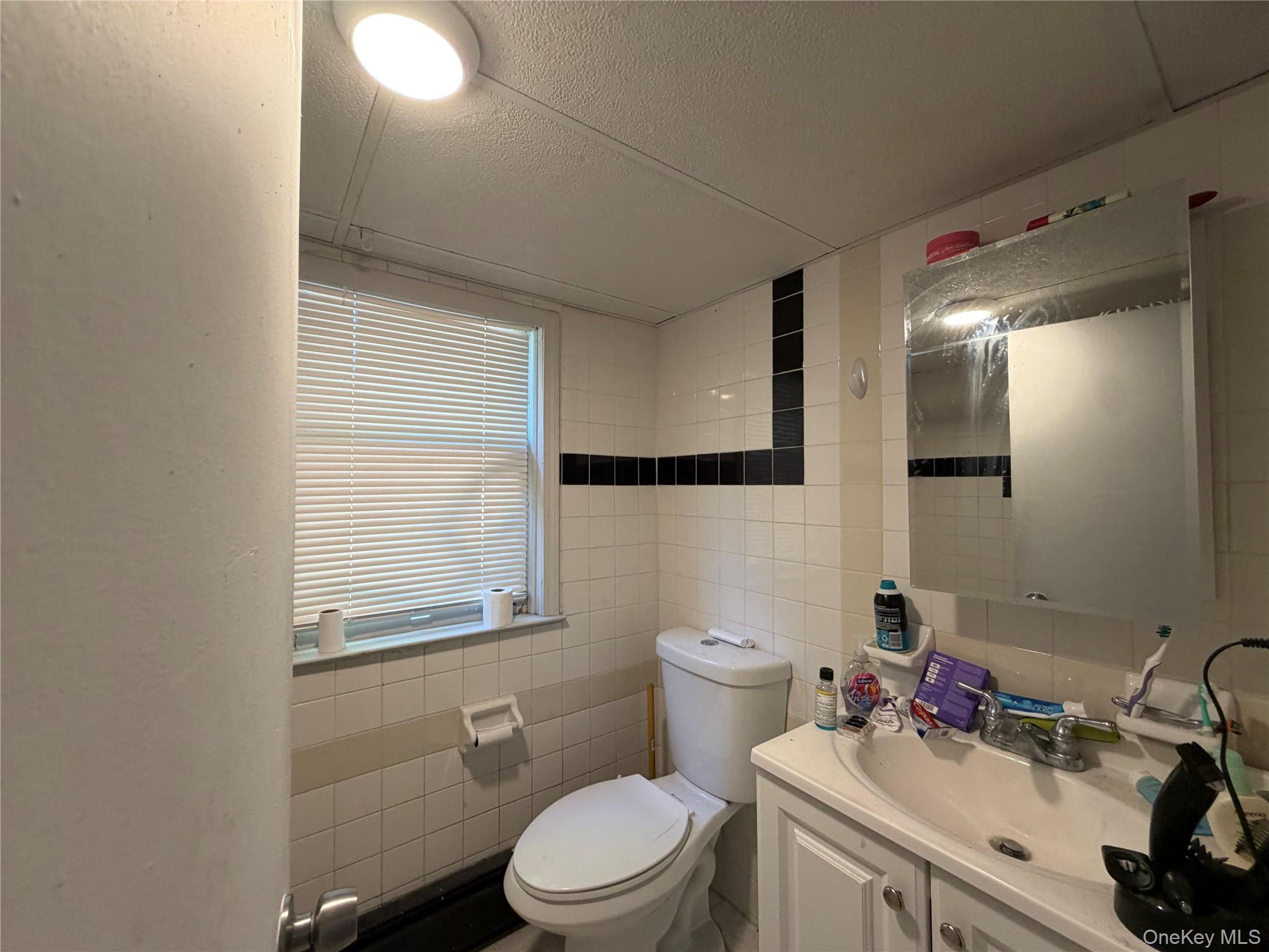 149 Fort Salonga Road Northport, NY 11768 - Photo 12 of 23 Bathroom featuring white wall tiling with black accent bands, a window with horizontal blinds, a white vanity with cabinet storage, and a wall-mounted mirror