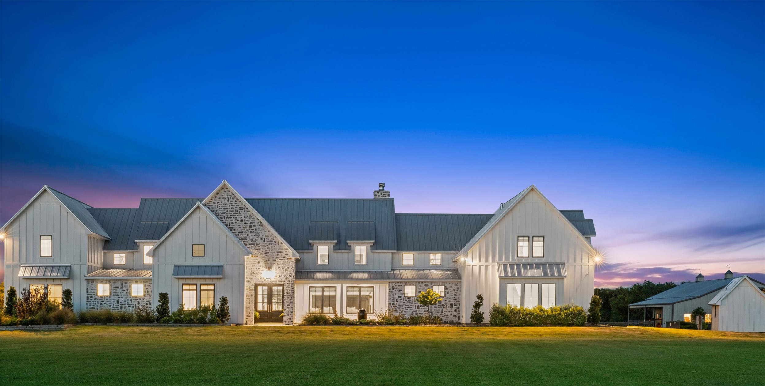 At twilight, the home’s stone façade, metal roofline, and glowing interior accents create a warm, refined presence that blends sophistication with rural charm.