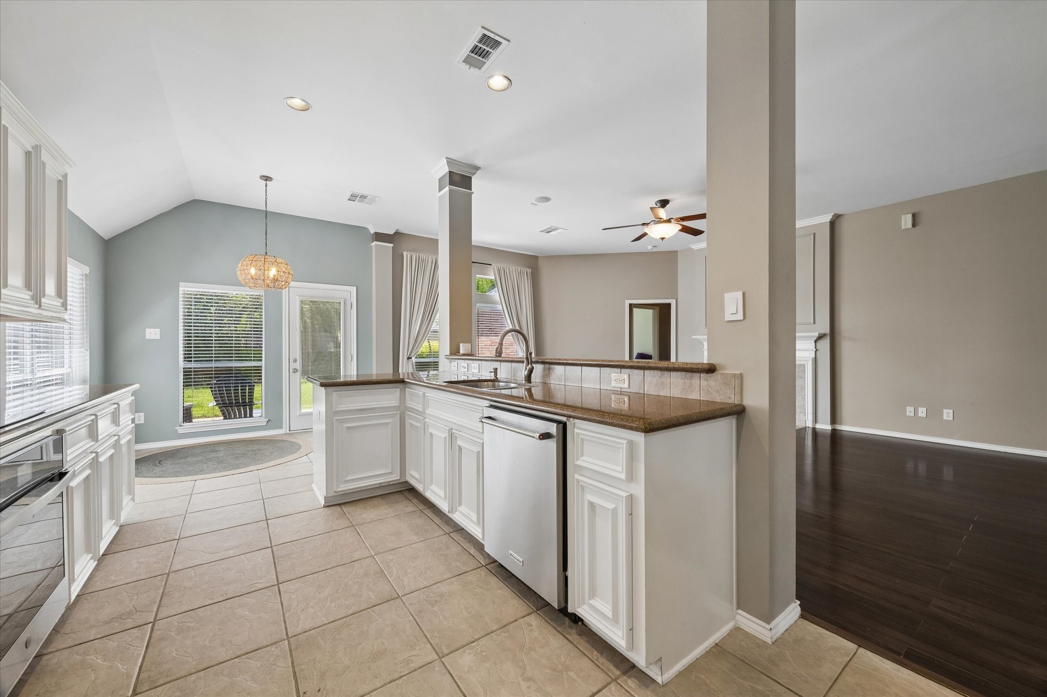 3118 Valley Court Manvel, TX 77578 - Photo 11 of 38 This photo showcases a spacious, open-concept kitchen with white cabinets, tiled floors, and a central island. It opens into a bright living area with a ceiling fan and large windows, offering a welcoming and airy atmosphere.
