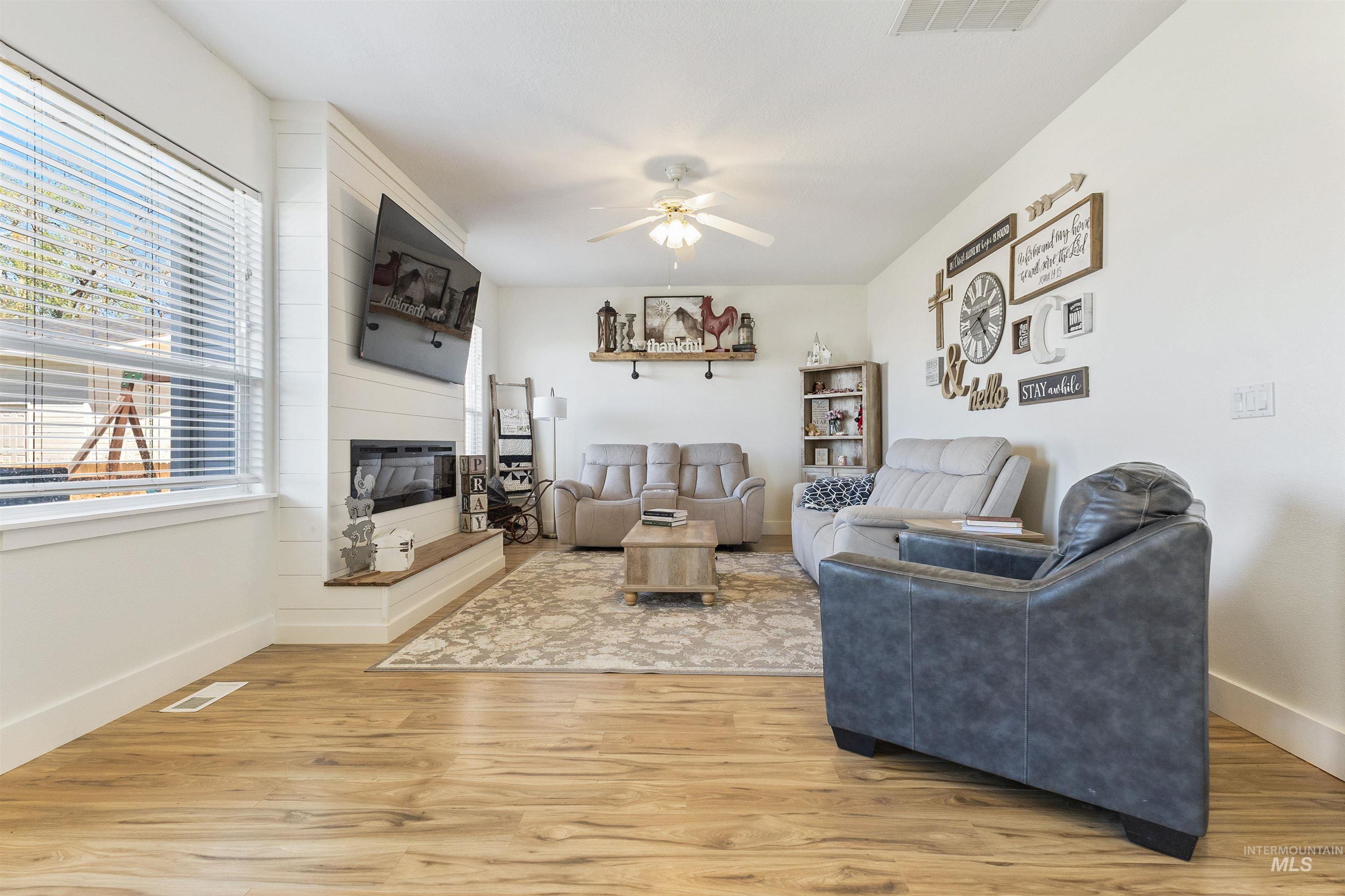 19876 Scotts Lane Caldwell, ID 83607 - Photo 28 of 44 Living area featuring wood-finish flooring, a contemporary electric fireplace with a white shiplap surround, a large window with blinds, a ceiling fan, and white baseboards