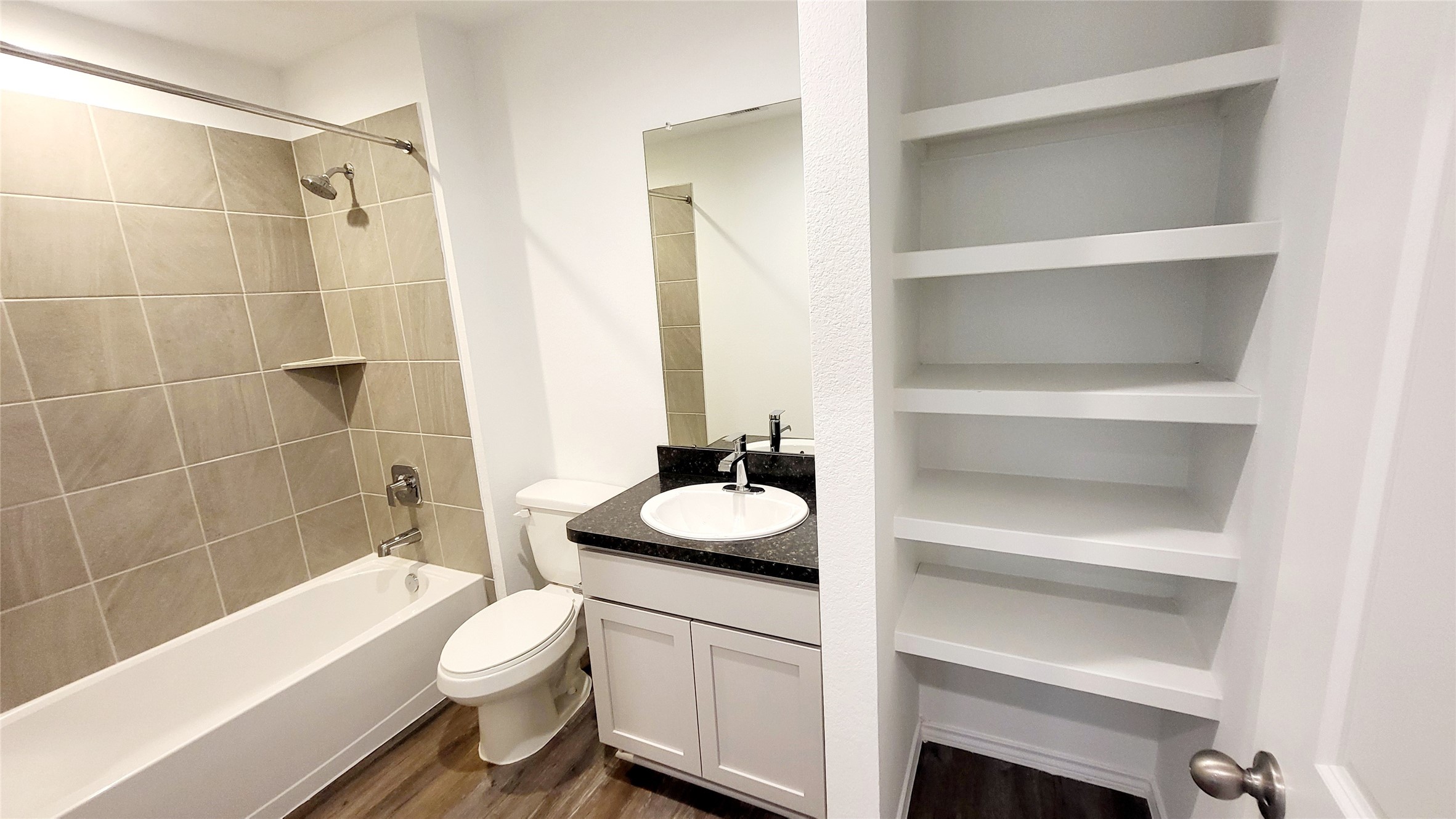 23835 Sunland Park Spring, TX 77373 - Photo 12 of 14 This is a modern bathroom featuring a bathtub with beige tiling, a toilet, a vanity with a black countertop, and built-in white shelves for storage.