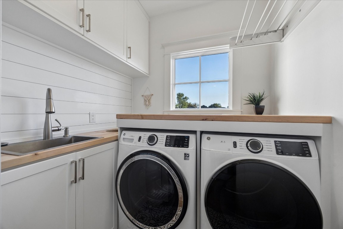 3535 Michaux Street Houston, TX 77009 - Photo 38 of 48 Here you will find a bright, modern laundry room featuring front-loading washer and dryer units. The space includes a sleek sink with a stainless steel faucet, white cabinetry, and a wooden countertop. A window provides natural light, and there are drying racks smartly positioned above. Barely visible at the back left corner are openings in a hinged counter section for dropping laundry into two bagged compartments of laundry that can be lifted out for simple sorting.