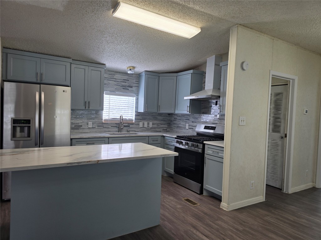 7628 Northview Lane Austin, TX 78724 - Photo 3 of 19 Kitchen featuring appliances with stainless steel finishes, gray cabinetry, a kitchen island, wall chimney exhaust hood, and a textured ceiling