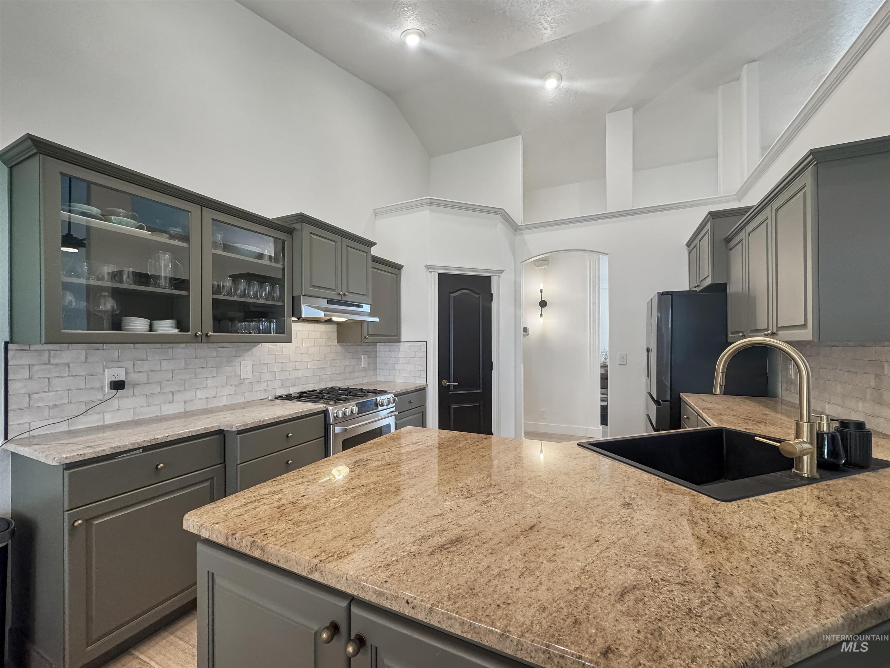 3917 North Waterloo Way Boise, ID 83713 - Photo 8 of 24 Kitchen features vaulted ceilings, recessed lighting, gray cabinetry with some glass-front doors, a subway tile backsplash, and a kitchen island with a dark sink and gold-finish faucet
