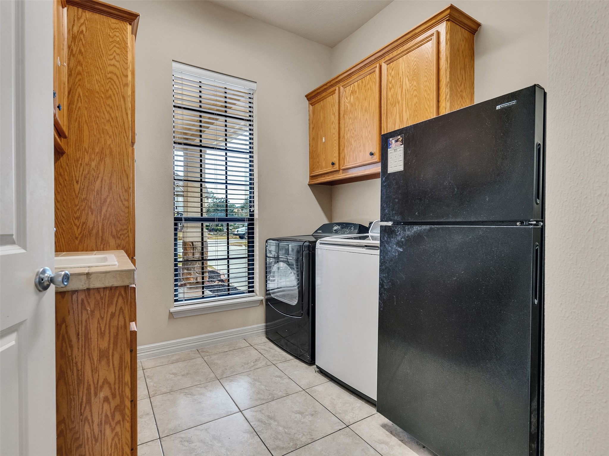 6914 Sandwedge Point Court Spring, TX 77389 - Photo 27 of 50 This is a compact laundry room featuring a black refrigerator, washer, and dryer. It has light-colored tile flooring, wooden cabinets for storage, and a window with blinds providing natural light.