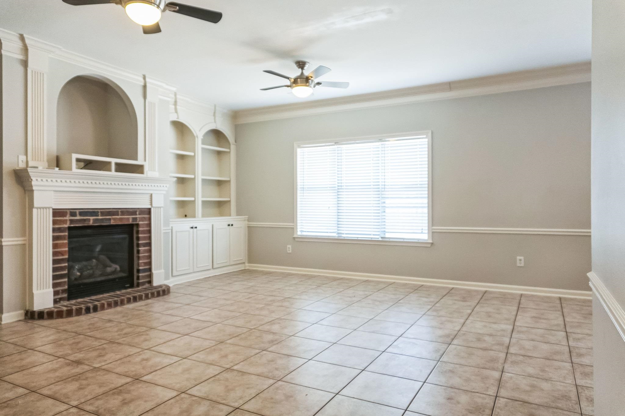 5225 Rockyford Road Bartlett, TN 38002 - Photo 6 of 18 Unfurnished living room with built in shelves, a brick fireplace, ceiling fan, ornamental molding, and light tile patterned flooring