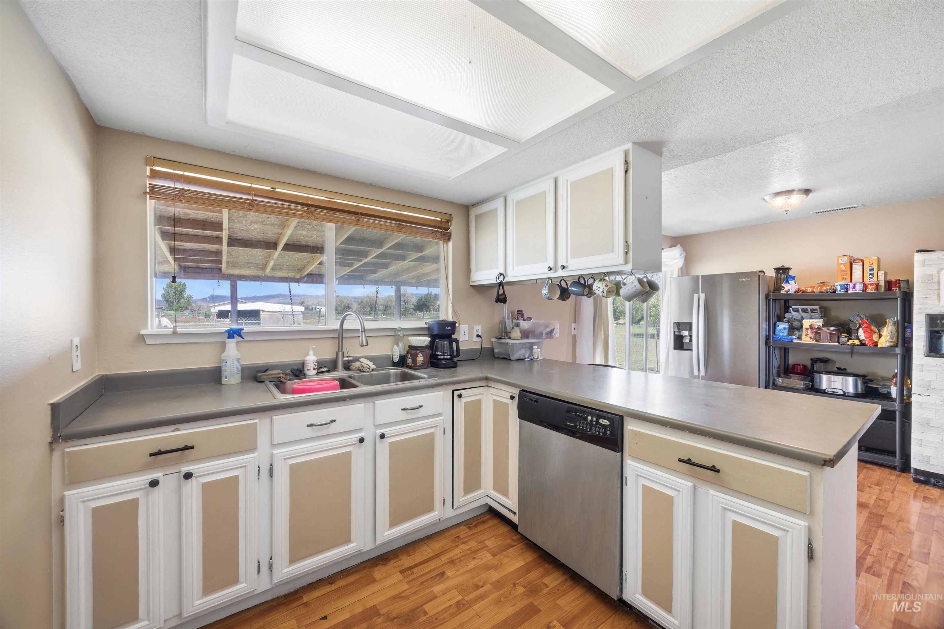 3044 Northeast Reservoir Road Mountain Home, ID 83647 - Photo 10 of 34 Kitchen featuring wood-finish flooring, L-shaped countertop, white cabinetry with tan accents, a stainless steel dishwasher, and a double basin sink beneath a window
