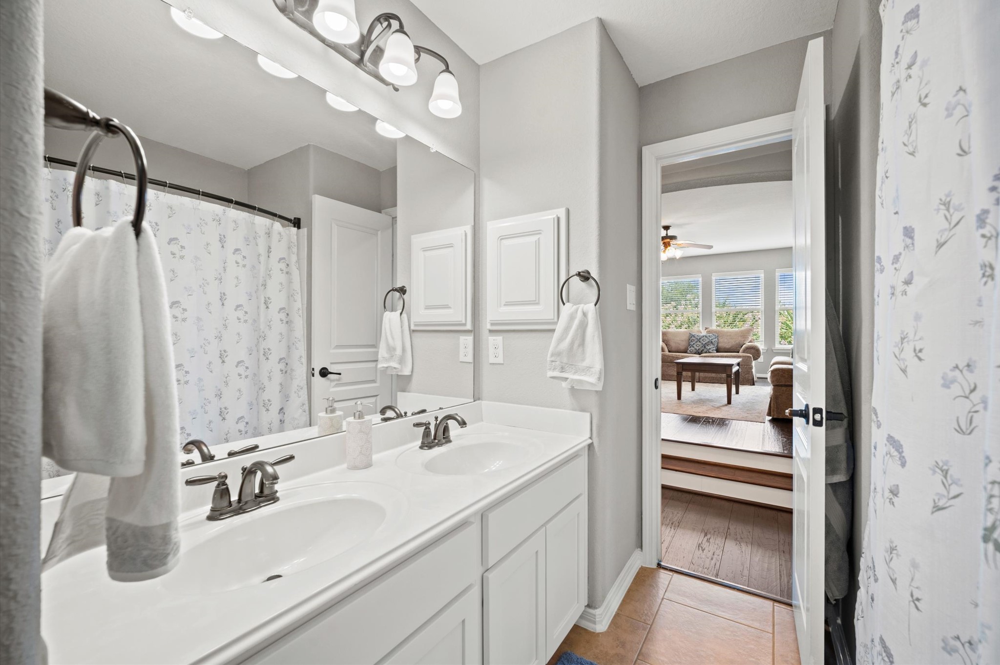 9507 Empress Crossing Drive Spring, TX 77379 - Photo 38 of 50 This photo showcases a bright bathroom with a double vanity, modern fixtures, and a shower with a floral curtain. It opens into a cozy living area with large windows, offering a view of the outdoors and ample natural light.