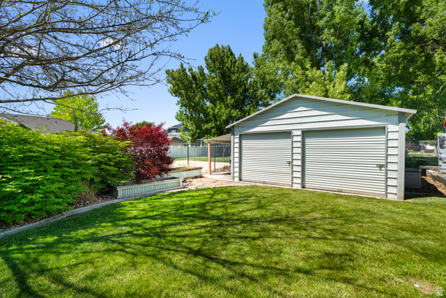 5727 South 3550th Street West Roy, UT 84067 - Photo 34 of 49 Expansive green lawn with a two-car shop featuring roll-up doors, surrounded by mature trees and shrubs