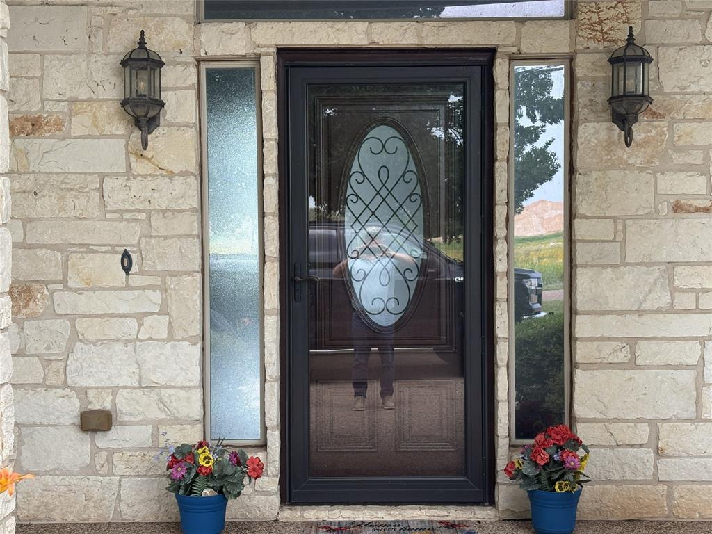 1170 Brazos Loop Gholson, TX 76705 - Photo 5 of 37 Stone facade entry featuring a dark storm door with decorative glass, two exterior sconce lights, and narrow sidelight windows