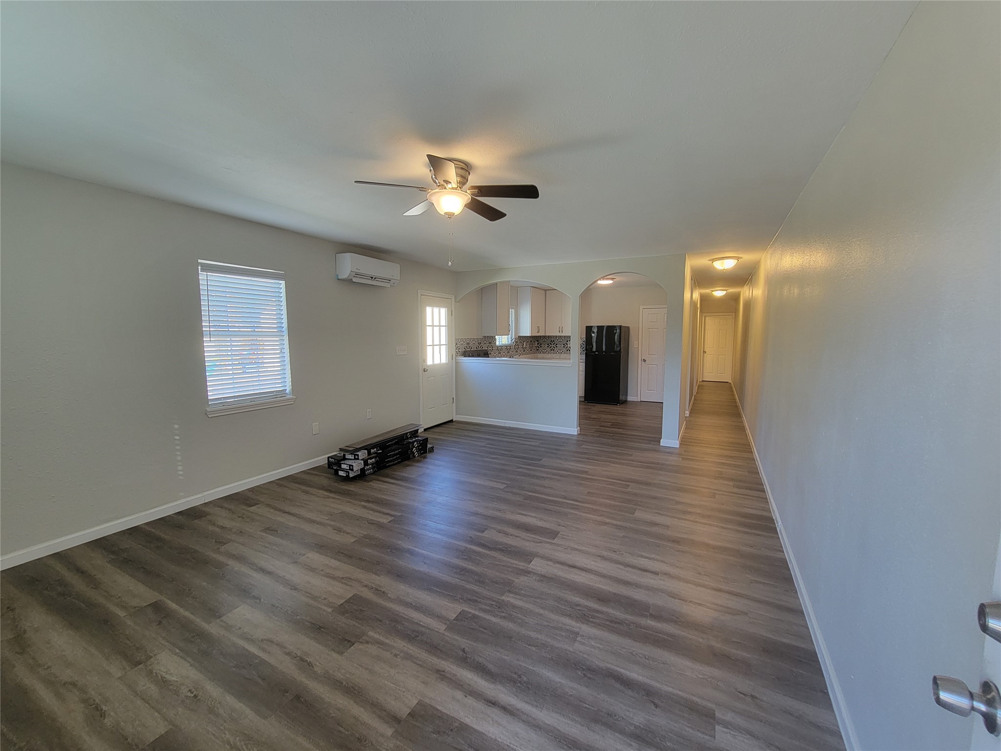 Spacious living area with modern vinyl plank flooring and neutral walls, featuring a ceiling fan and ample natural light from multiple windows. The open layout leads to a kitchen with an arched entrance and a view of the hallway and doorways.