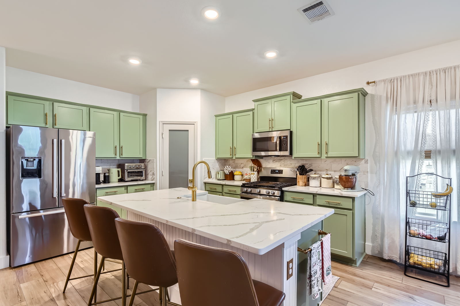 Kitchen with stainless steel appliances, green cabinets, backsplash, light stone counters, and a center island with sink