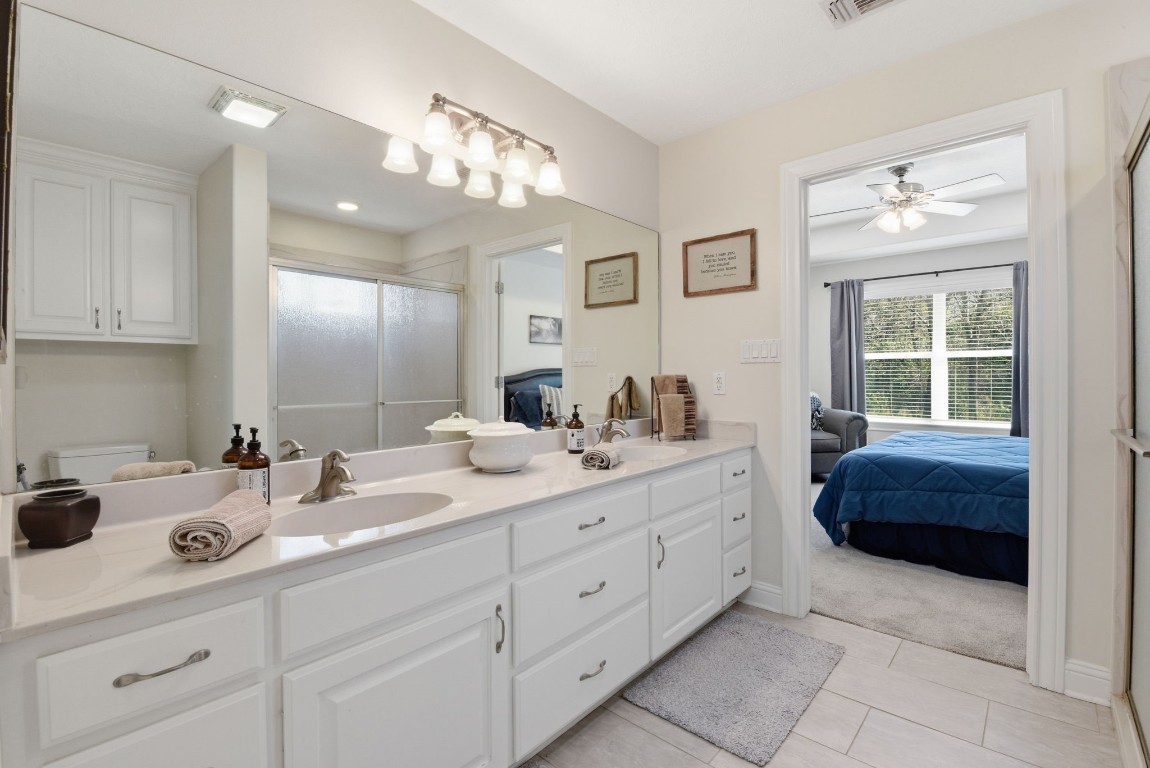 232 North Dixie Street Brenham, TX 77833 - Photo 29 of 41 This photo showcases a bright, modern bathroom with a dual-sink vanity, ample storage, and a large mirror. The room connects to a cozy bedroom with large windows with views of lush greenery.