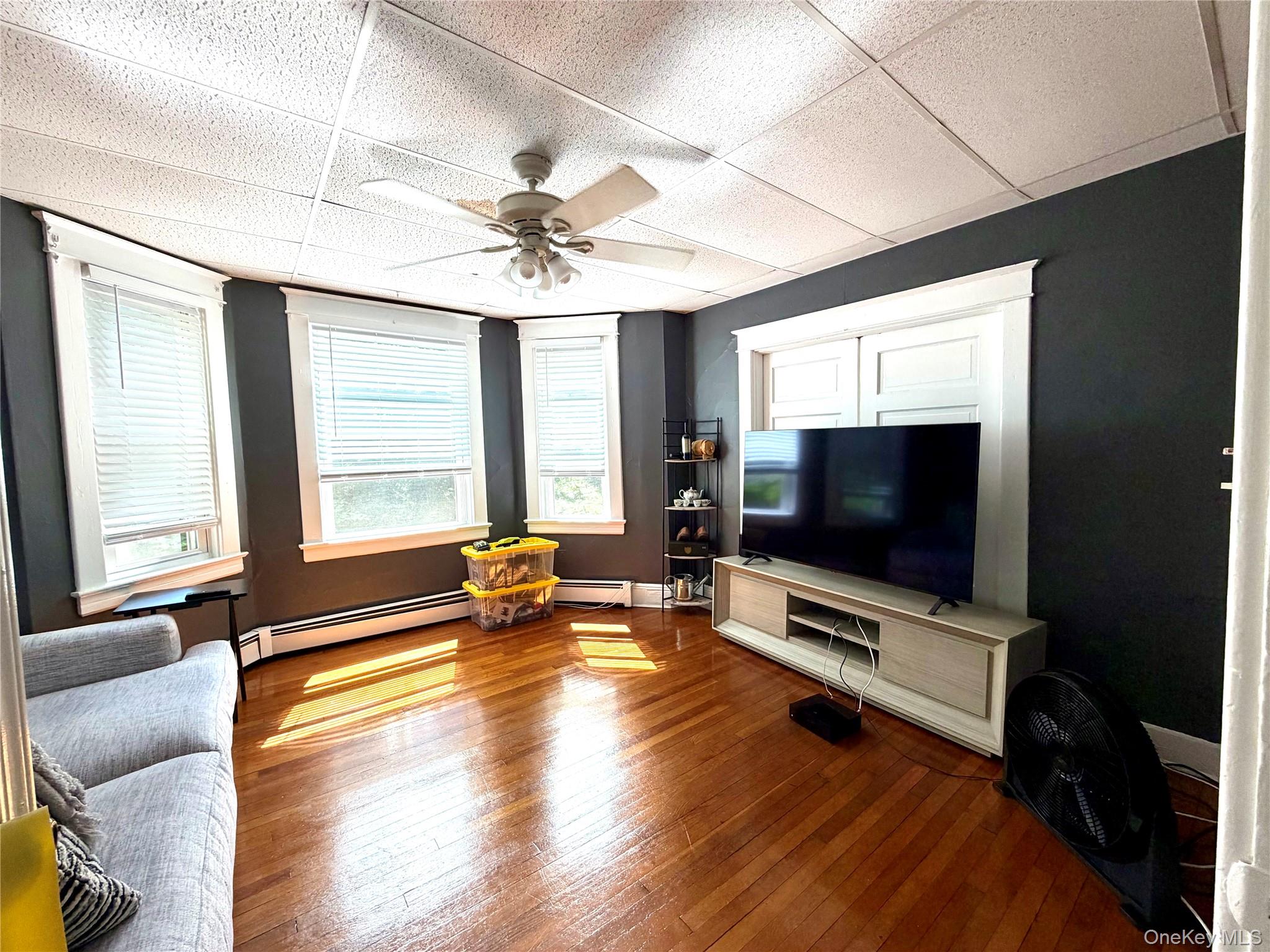 Living area featuring wood-finish flooring, a ceiling fan with integrated lighting, and multiple windows with white blinds
