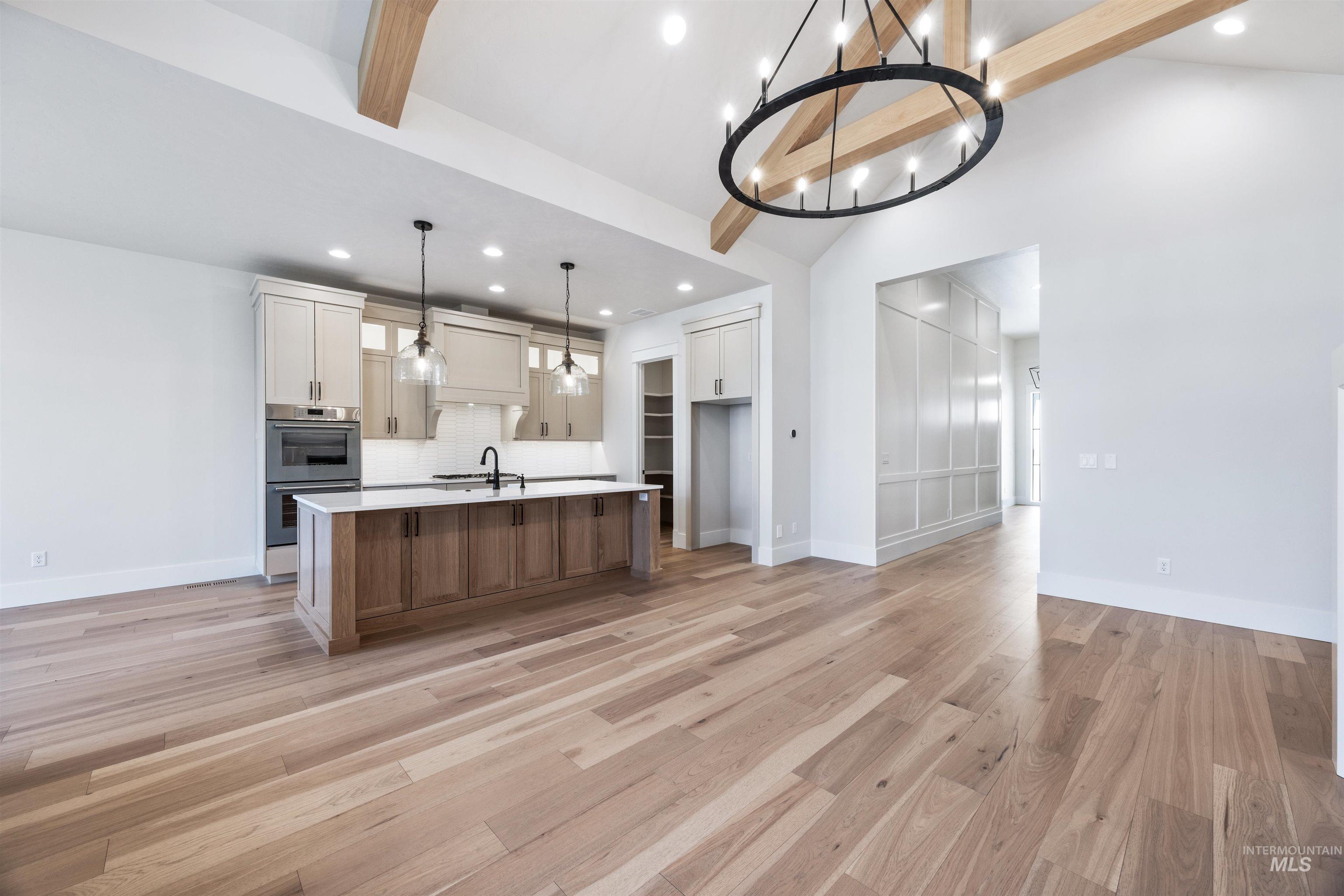 904 East Escalation Street Meridian, ID 83642 - Photo 11 of 36 Open-concept kitchen and living area featuring wood-finish flooring, a large kitchen island with a white countertop, built-in double ovens, a striking black chandelier, and exposed ceiling beams