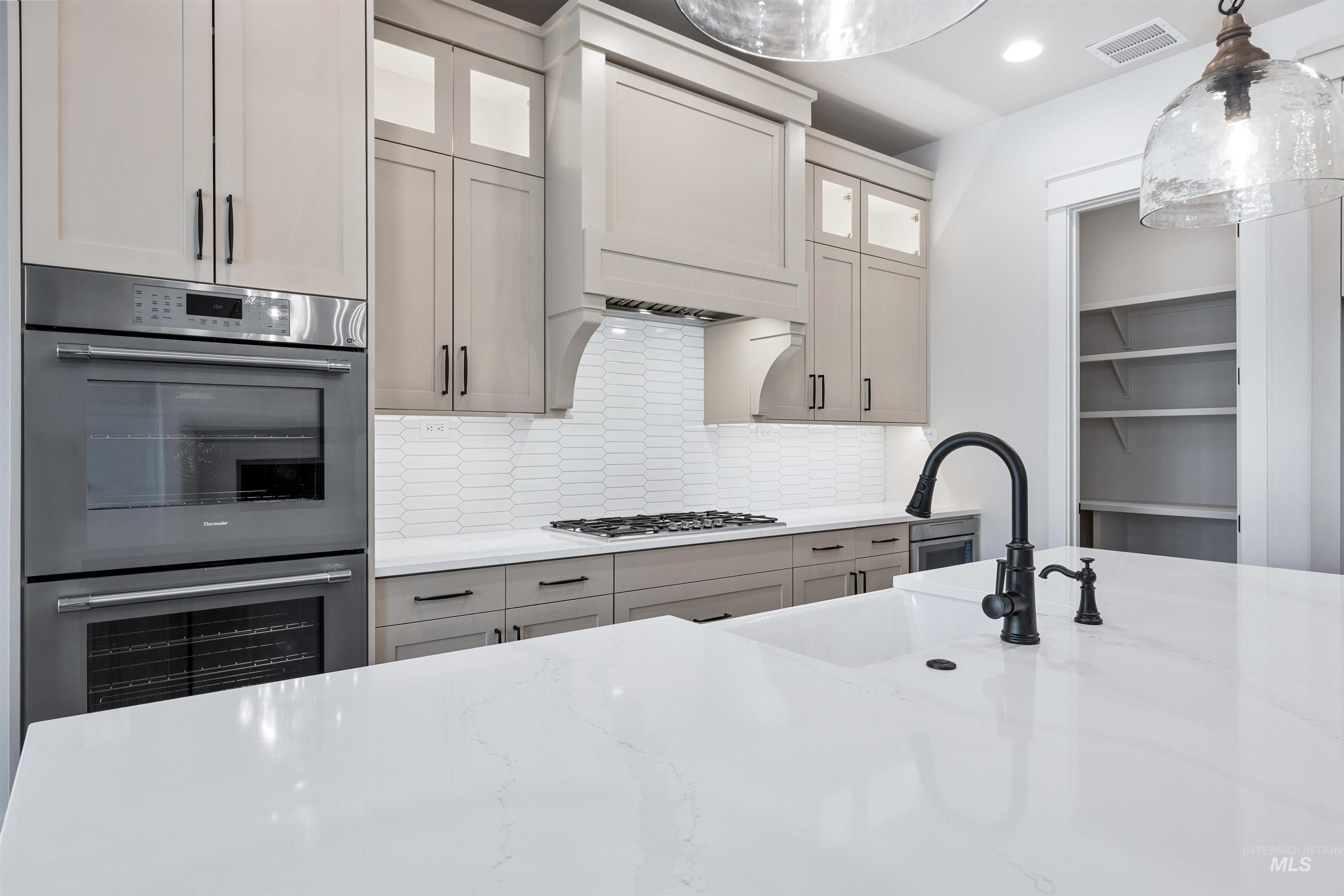 904 East Escalation Street Meridian, ID 83642 - Photo 17 of 36 Modern kitchen featuring light cabinetry, stainless steel double ovens, a gas cooktop, a white picket tile backsplash, and an island with a built-in sink and matte black faucet