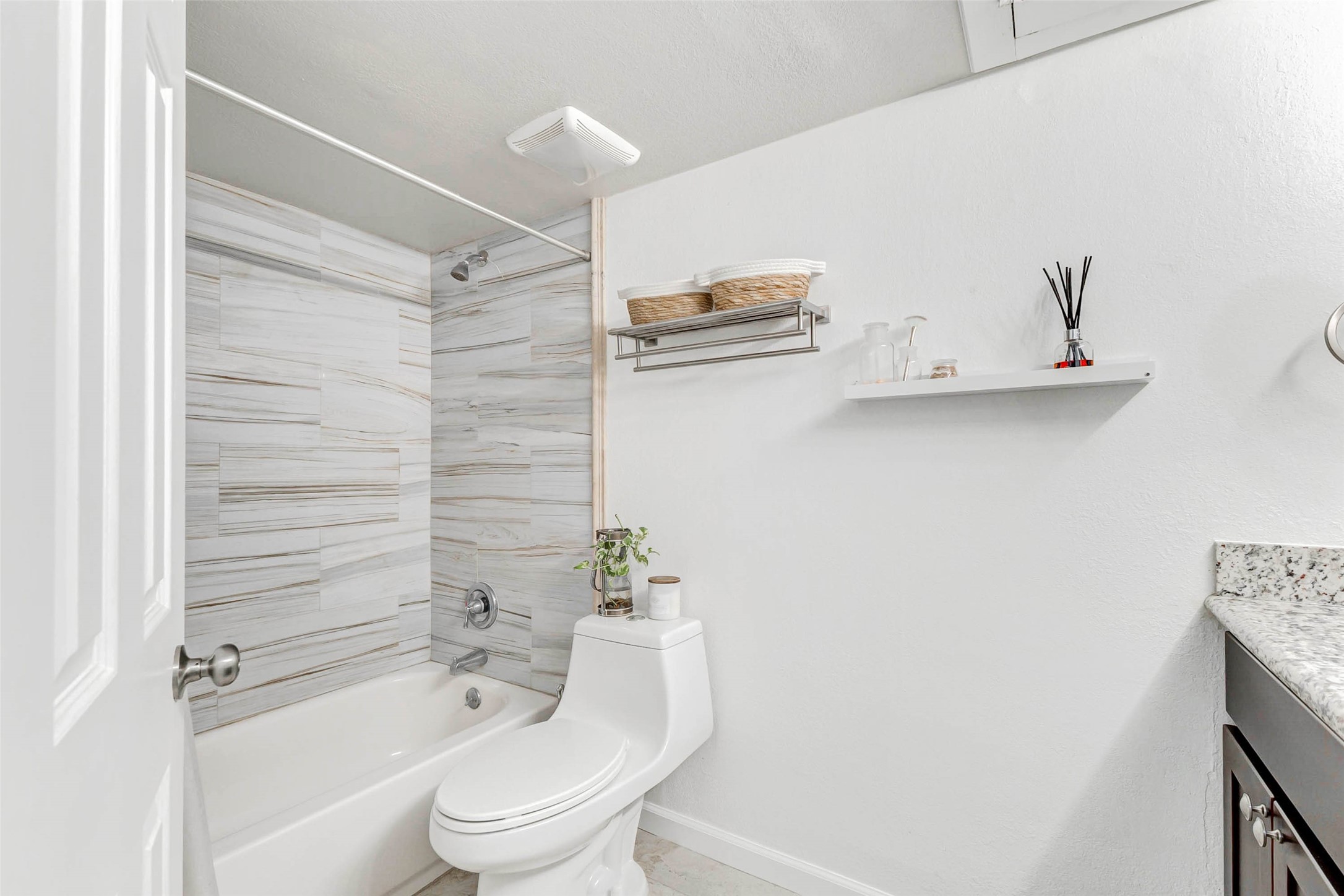 3928 West Alabama Street, Unit 4 Houston, TX 77027 - Photo 21 of 33 This bathroom features a modern design with a sleek white bathtub and shower combo, accented by stylish gray tile. It includes a contemporary toilet, open shelving for storage, and a granite countertop with a dark wood vanity. The space is bright and clean, offering a fresh and inviting atmosphere.