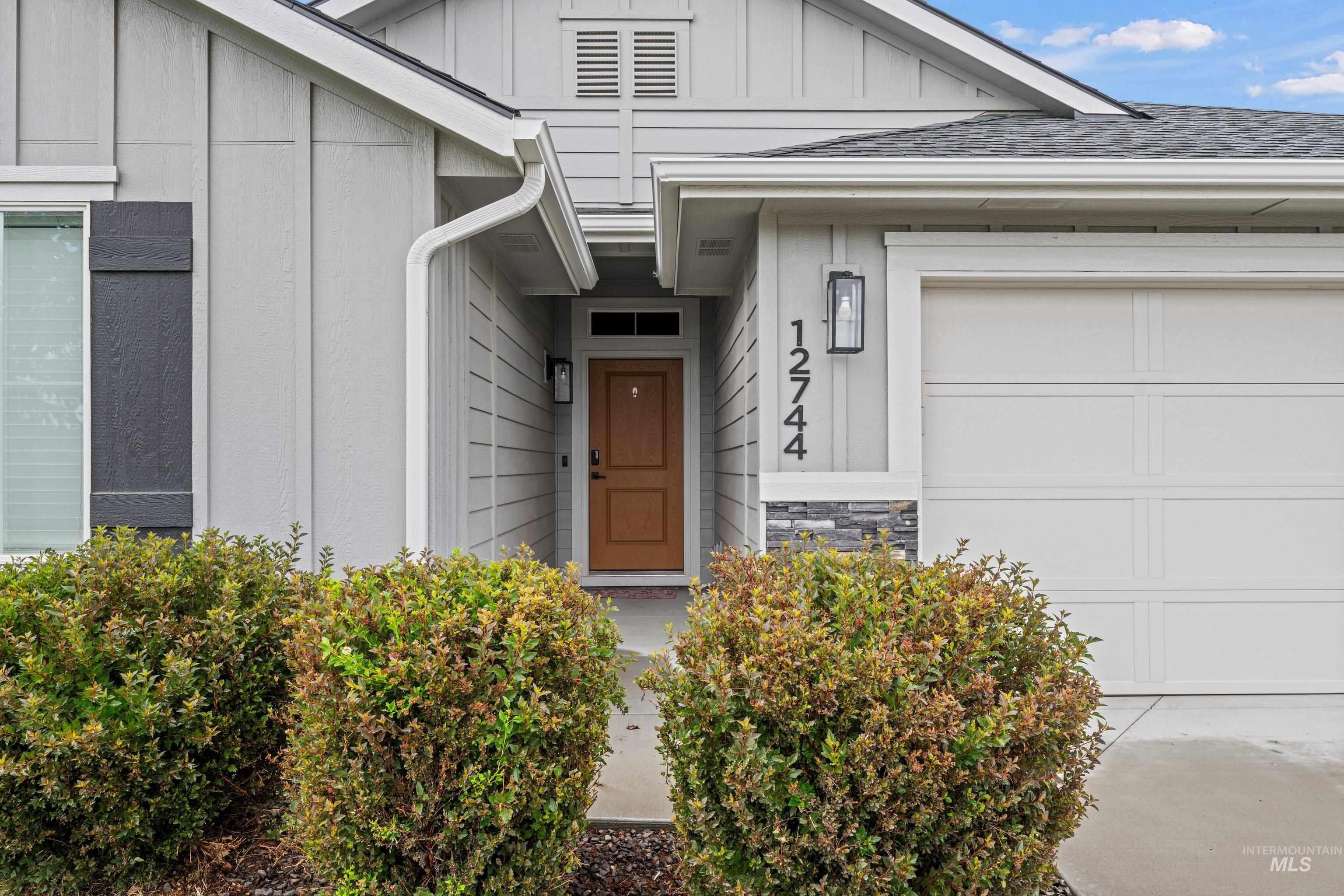 12744 West Fig Street Boise, ID 83713 - Photo 4 of 34 Contemporary exterior featuring light gray siding, a tan front entry door, a white garage door, stacked stone accents, and dark wood-tone shutters