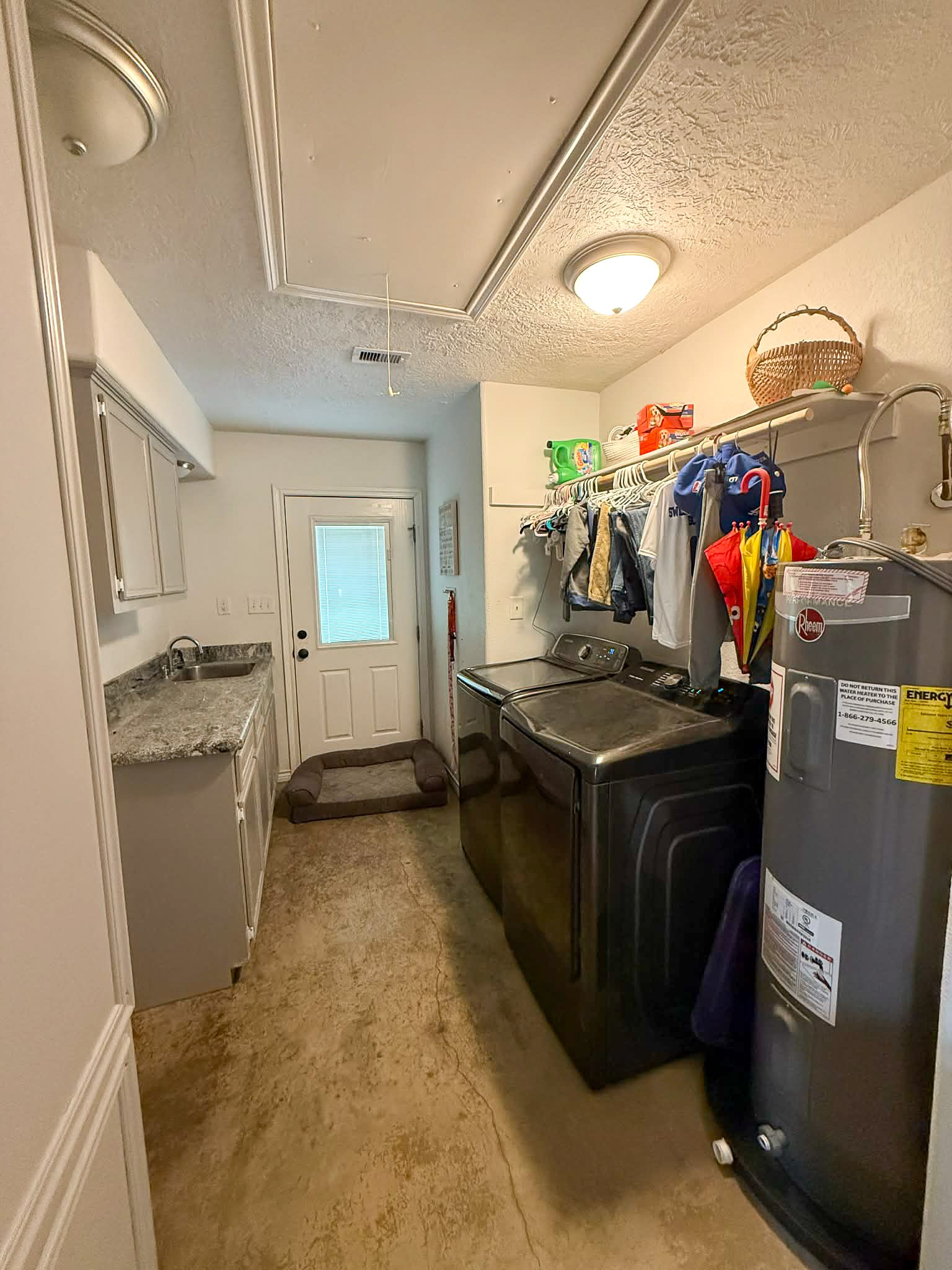 107 County Road 409 Sweeny, TX 77480 - Photo 12 of 20 This photo shows a functional laundry room with a washer, dryer, and water heater. It features a countertop with a sink, overhead cabinets for storage, and a clothing rod. The room has a door leading outside, providing convenient access.