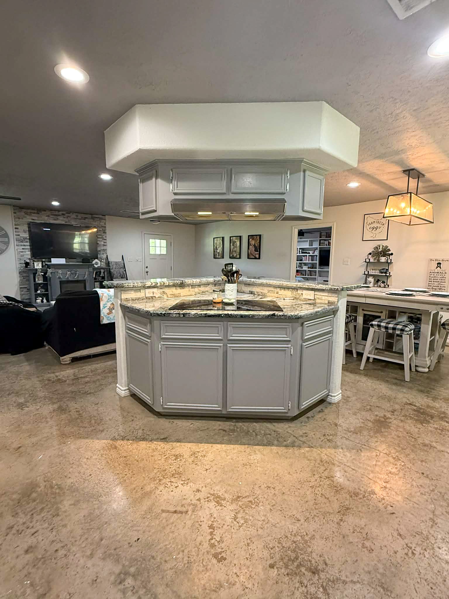 107 County Road 409 Sweeny, TX 77480 - Photo 6 of 20 This photo showcases a spacious open-concept kitchen and living area with a central island featuring quartz countertops. The kitchen has overhead cabinets and modern lighting, while the adjacent living area includes a cozy seating space with a fireplace and TV. The flooring is polished, adding a sleek look to the space.