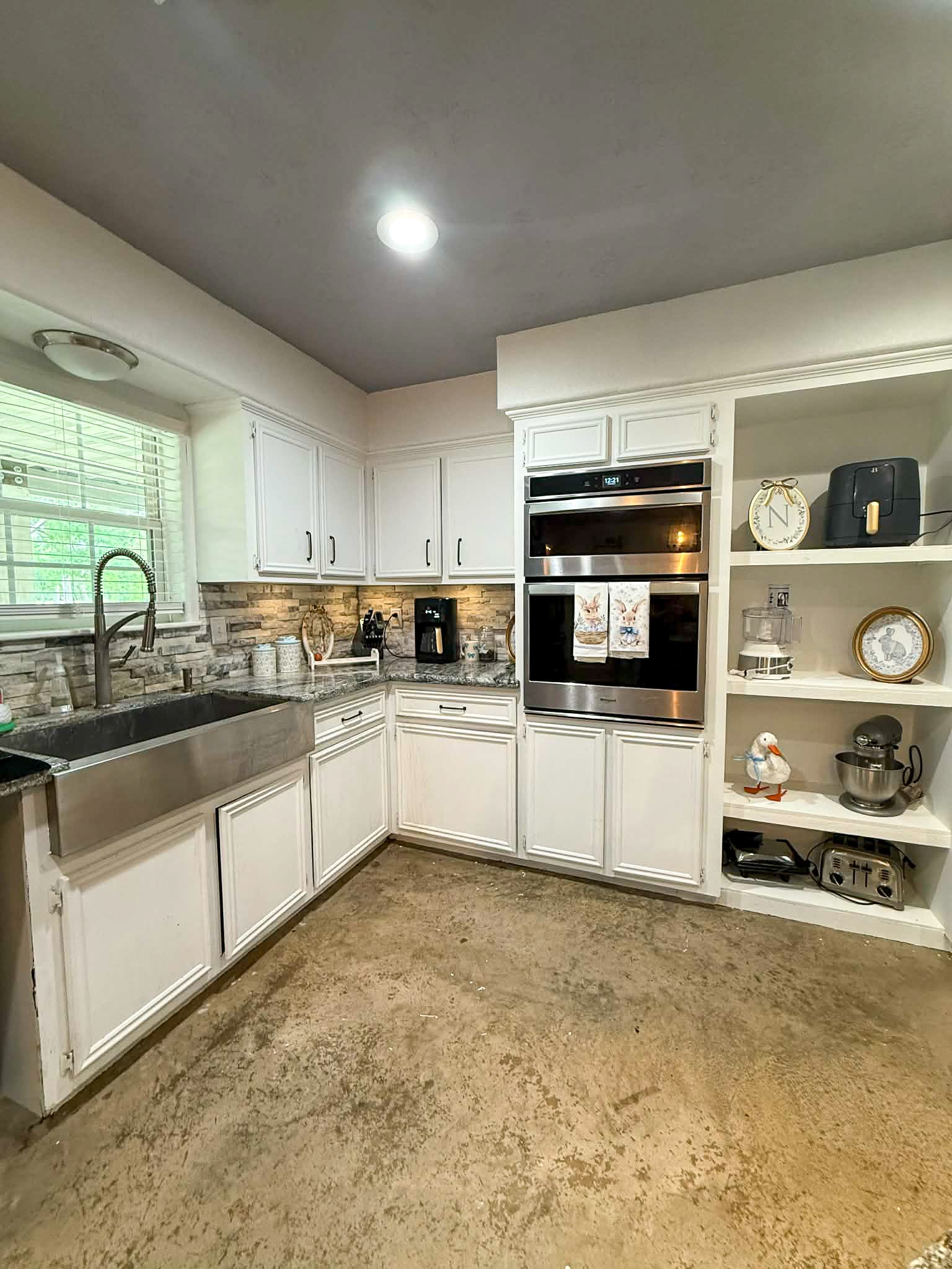 107 County Road 409 Sweeny, TX 77480 - Photo 7 of 20 This kitchen features a modern design with white cabinetry and stainless steel appliances, including a double oven. It has a farmhouse sink, a stone backsplash, and ample open shelving for storage. The space is well-lit with a large window and overhead lighting.